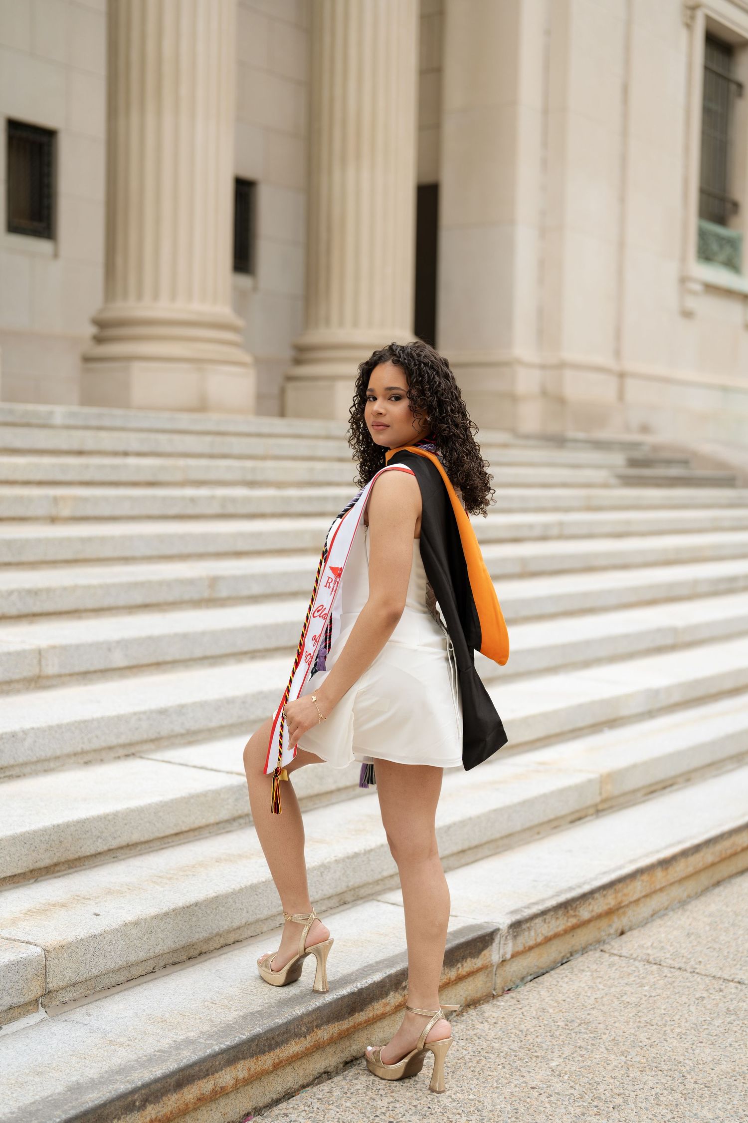 Young woman in white dress with graduation regalia poses on university building steps.