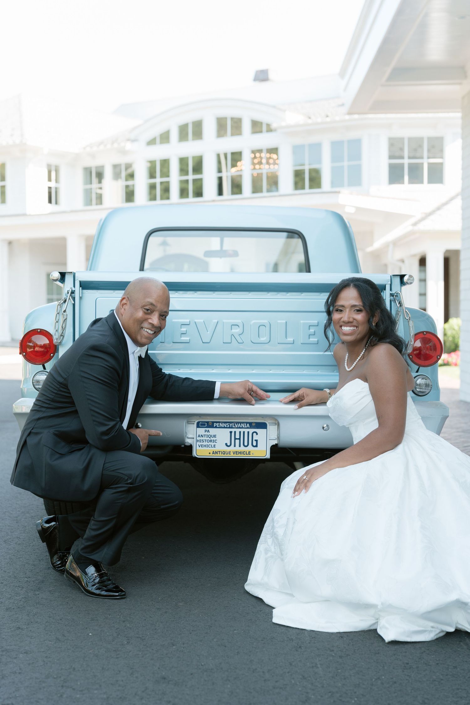 Smiling bride and groom pose beside a vintage light blue Chevrolet pickup truck with custom JHUG license plate.