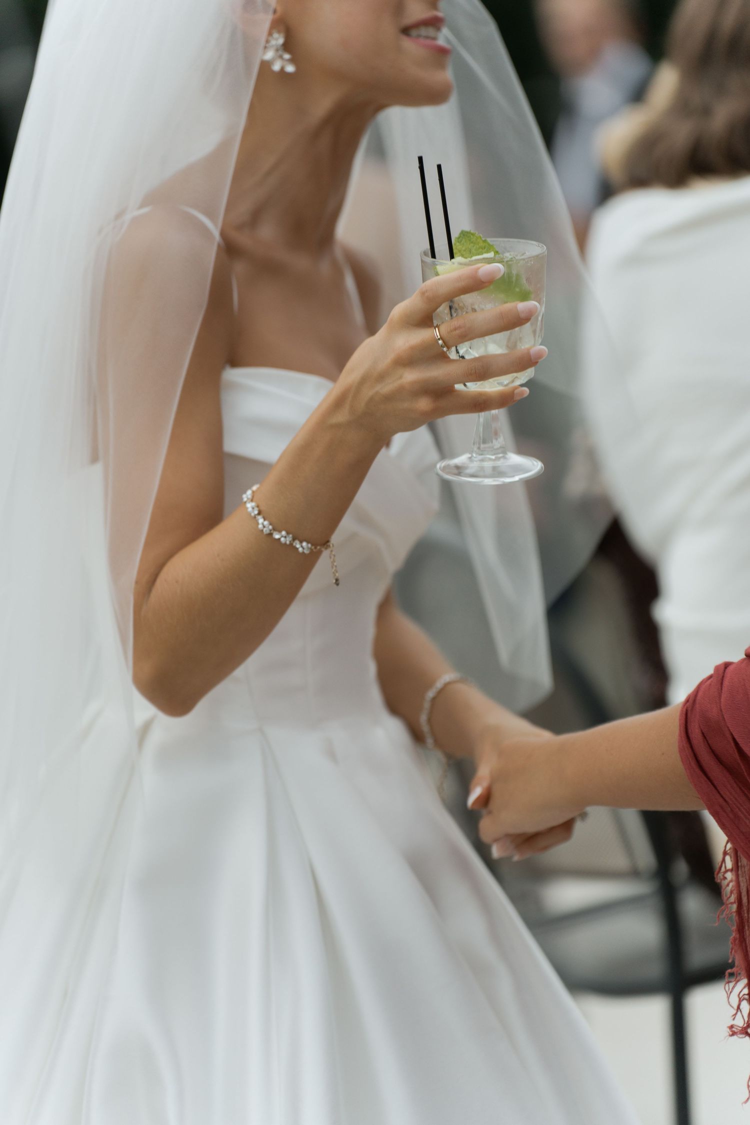 Smiling bride in white gown and veil holds a cocktail with lime garnish at a wedding reception.
