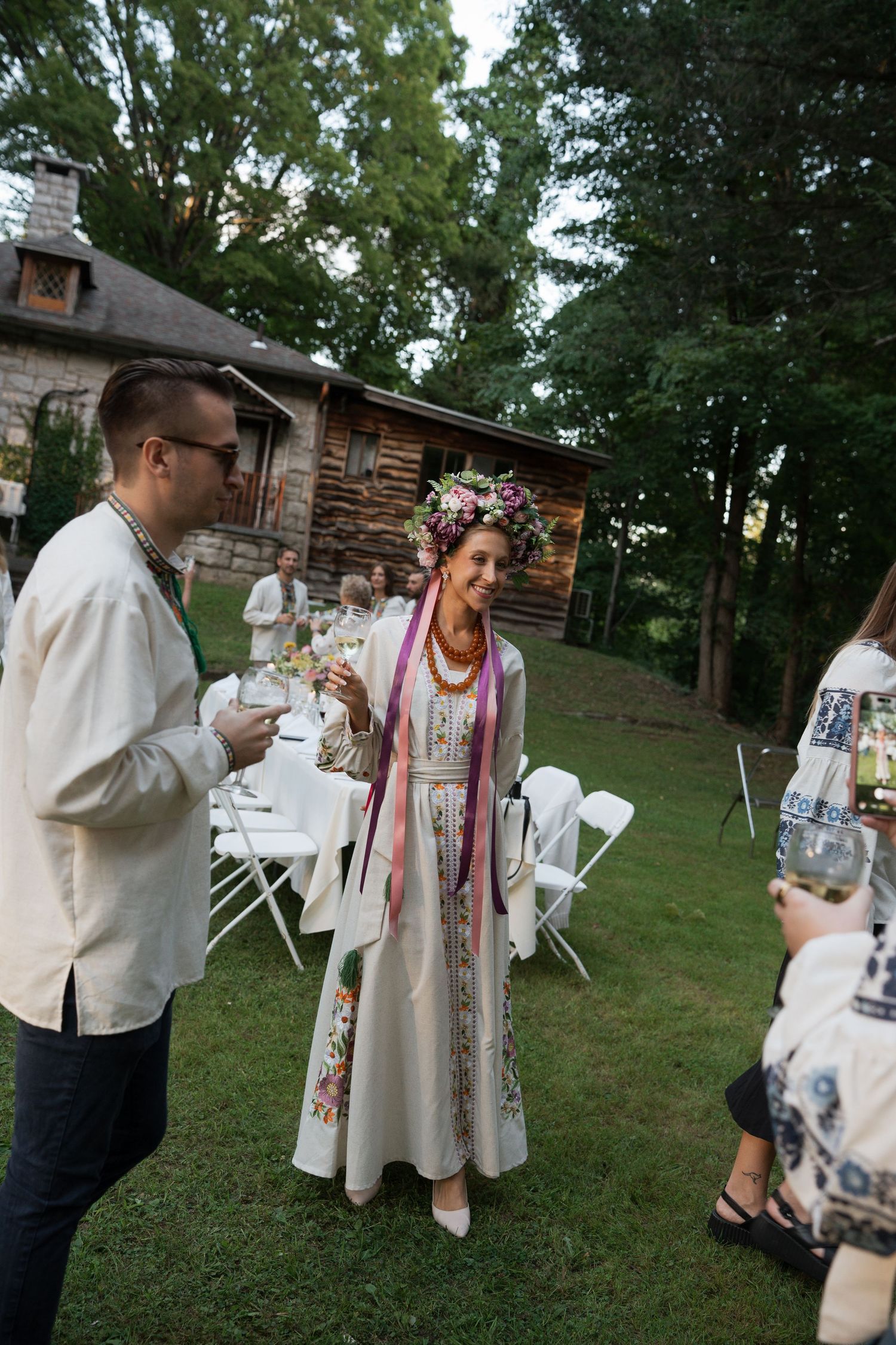 Woman in embroidered Ukrainian dress and floral crown at outdoor wedding reception with guests.