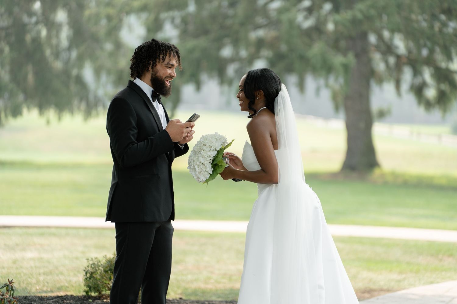 A groom in a black tuxedo reads vows on his phone to his bride in a white wedding gown outdoors.