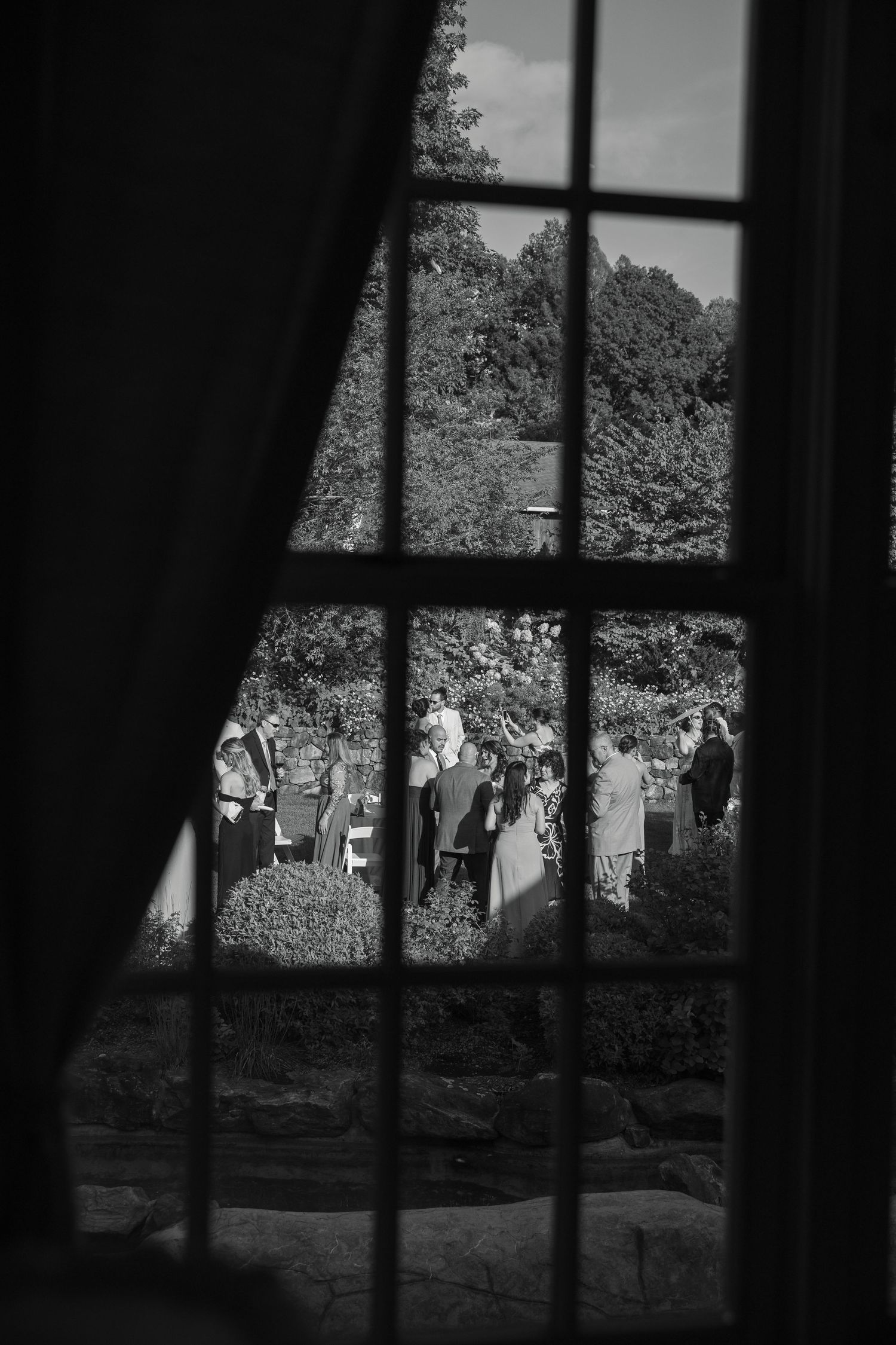Black and white view through window of wedding guests gathered in lush garden outdoor reception.