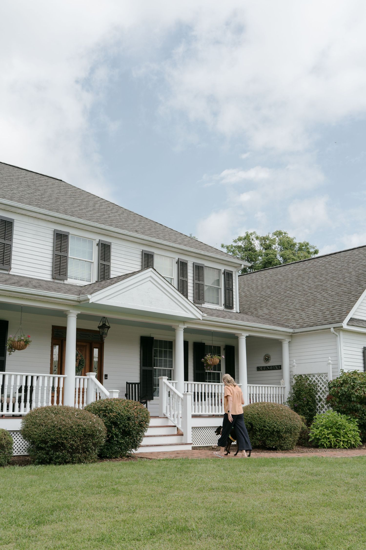Two women walk in front of a charming white colonial home with a wraparound porch and manicured shrubs.