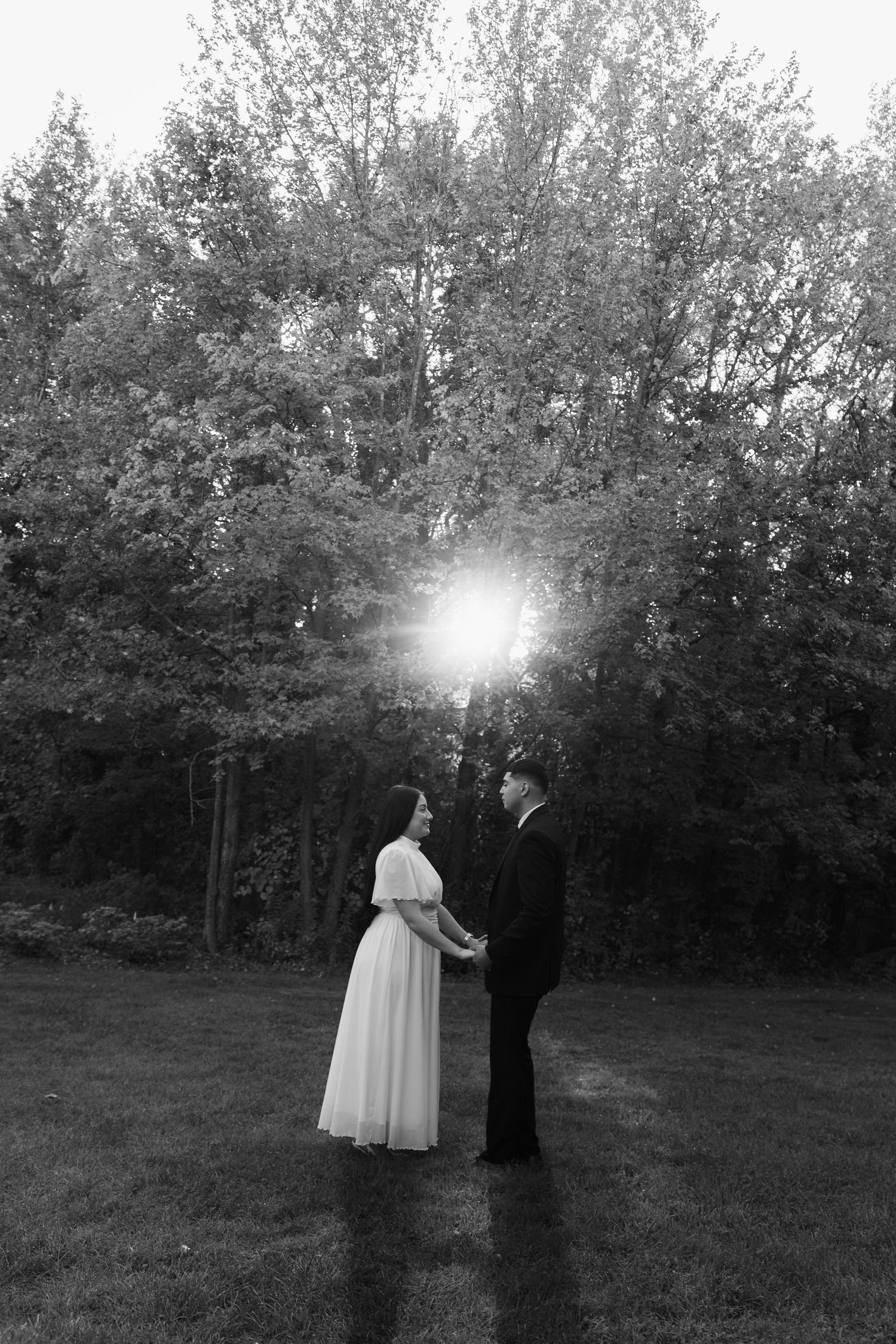 A couple stands together in formal attire as sunlight streams through trees behind them.