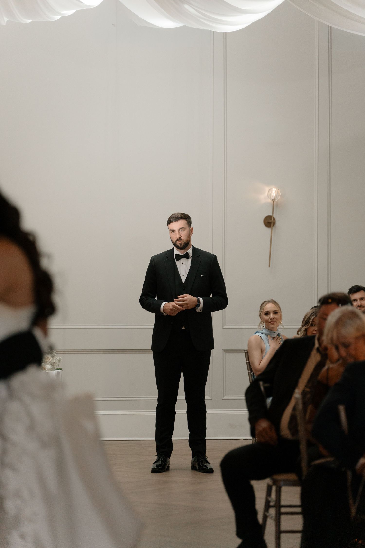 Groom in black tuxedo stands at altar awaiting bride during indoor wedding ceremony.