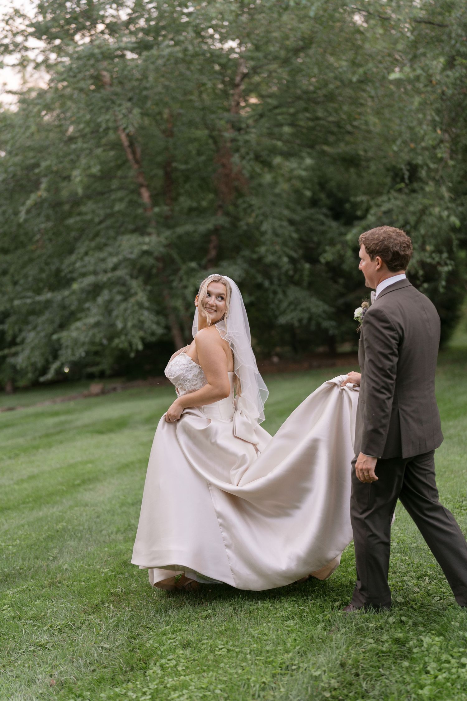 Bride in white gown and veil smiles as groom lifts her dress train on lush green lawn at outdoor wedding.