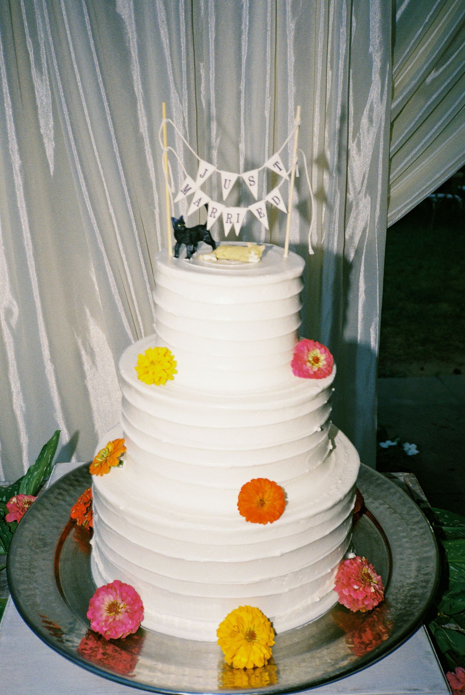 A white tiered wedding cake decorated with colorful flowers and a banner topper.