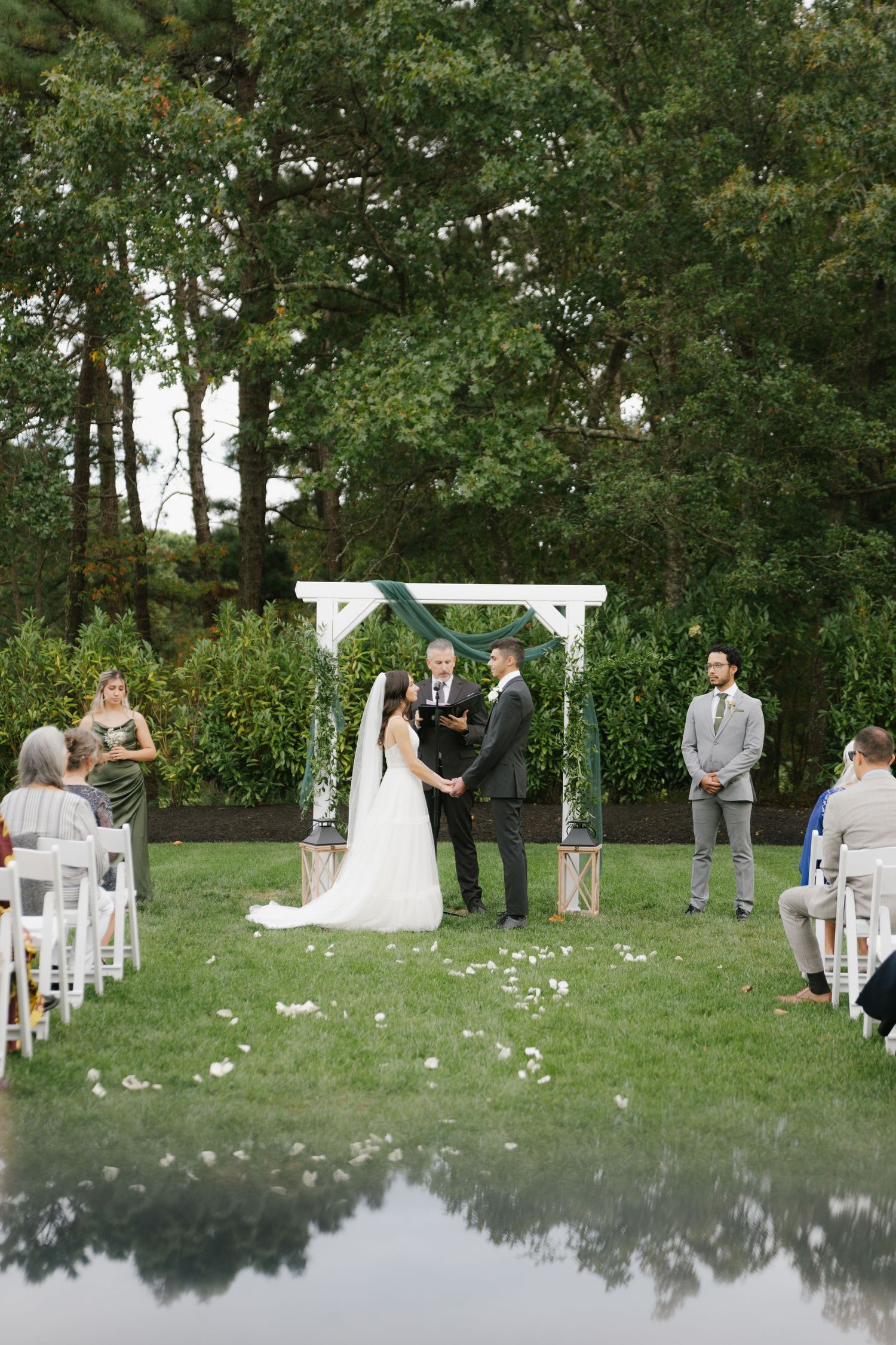 A wedding ceremony in progress on a manicured lawn with white chairs and a decorated arch against a backdrop of tall trees.