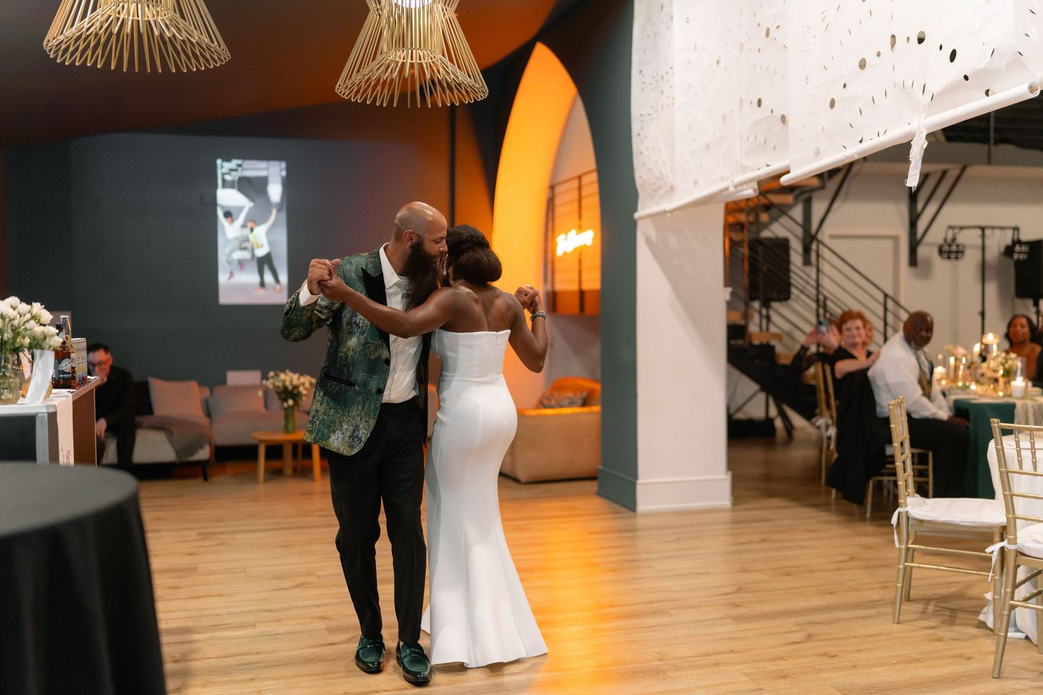 A couple dances together in a warmly lit event space with hardwood floors and pendant lighting.