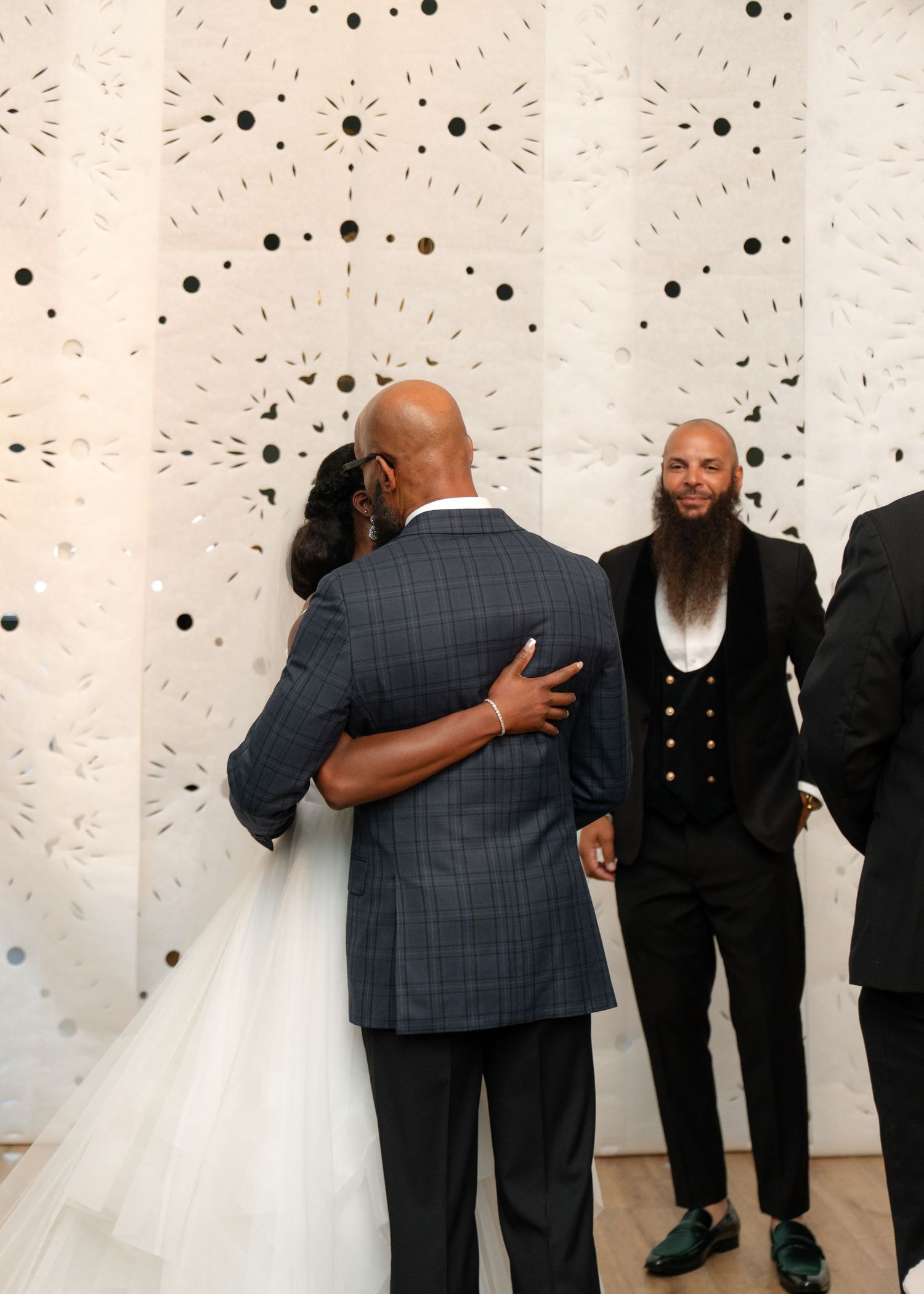 Wedding party sharing embraces and congratulations in front of a decorative dotted wall.