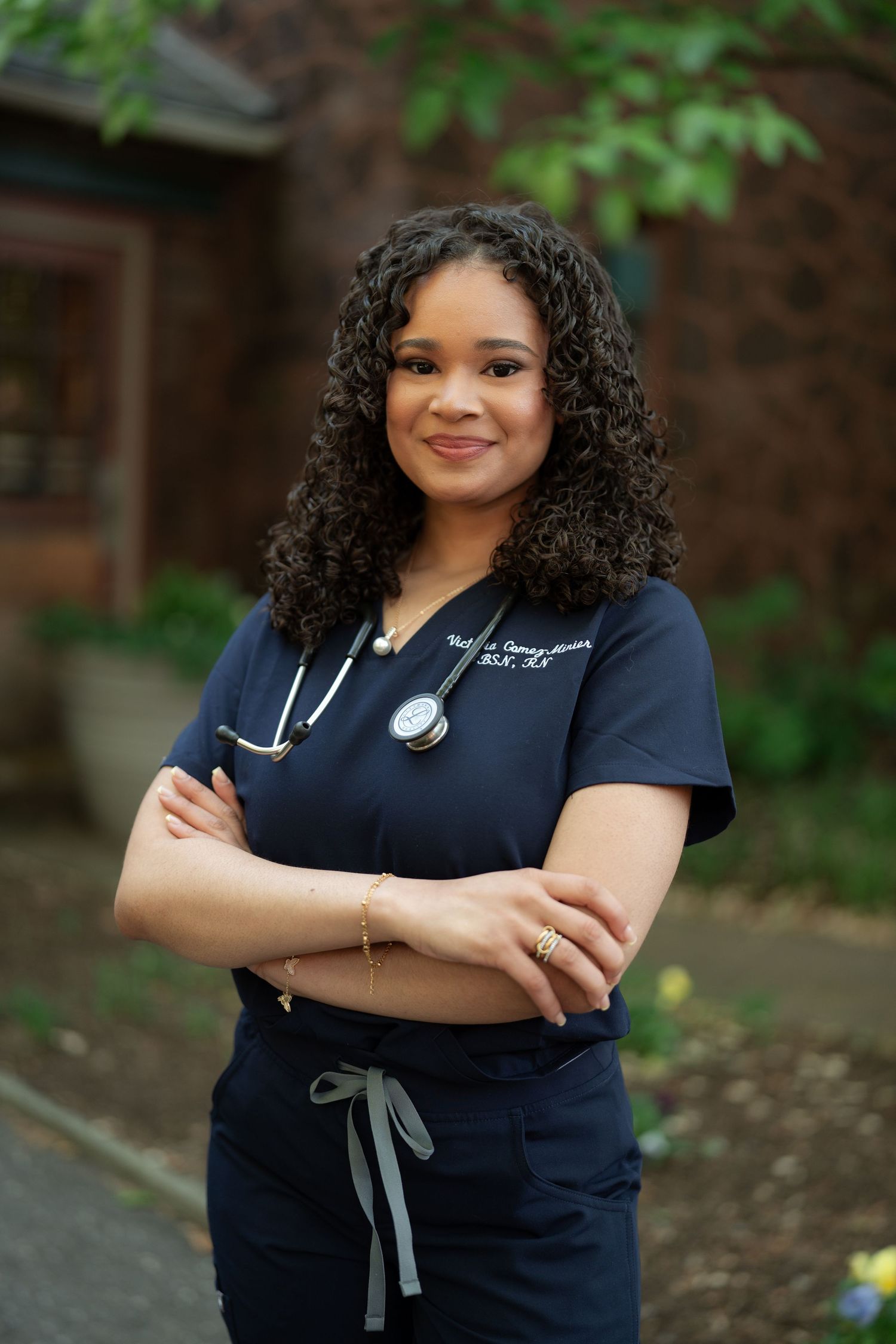 Female nurse in navy scrubs with stethoscope standing arms crossed outdoors with greenery background.