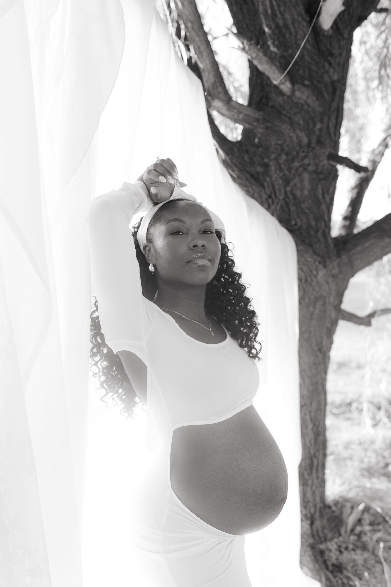 Black and white maternity photo of a pregnant woman in a white dress posing gracefully outdoors near a tree.