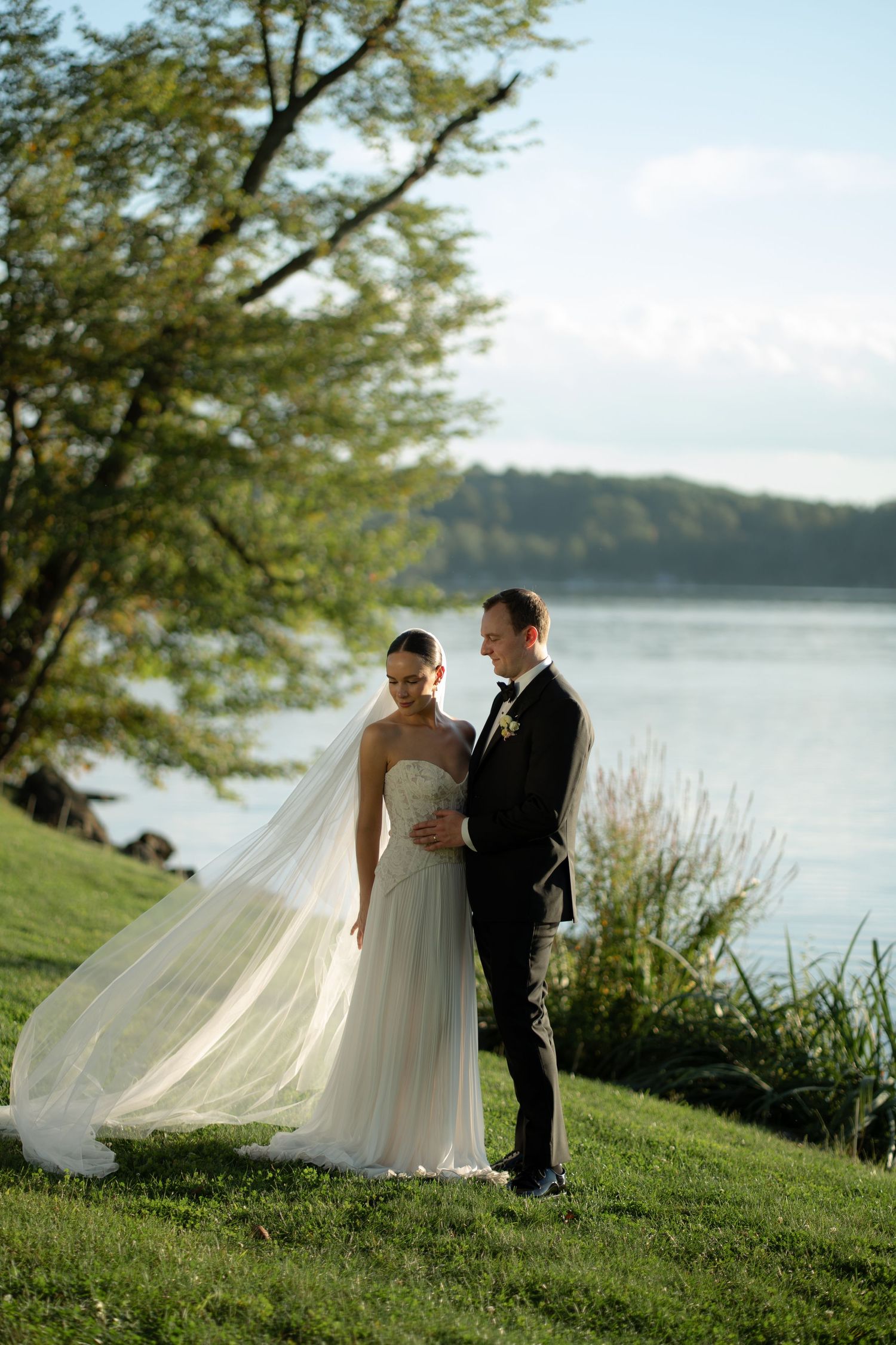 Bride and groom stand by a serene lakeside on green grass, her veil flowing in the breeze, surrounded by lush trees.