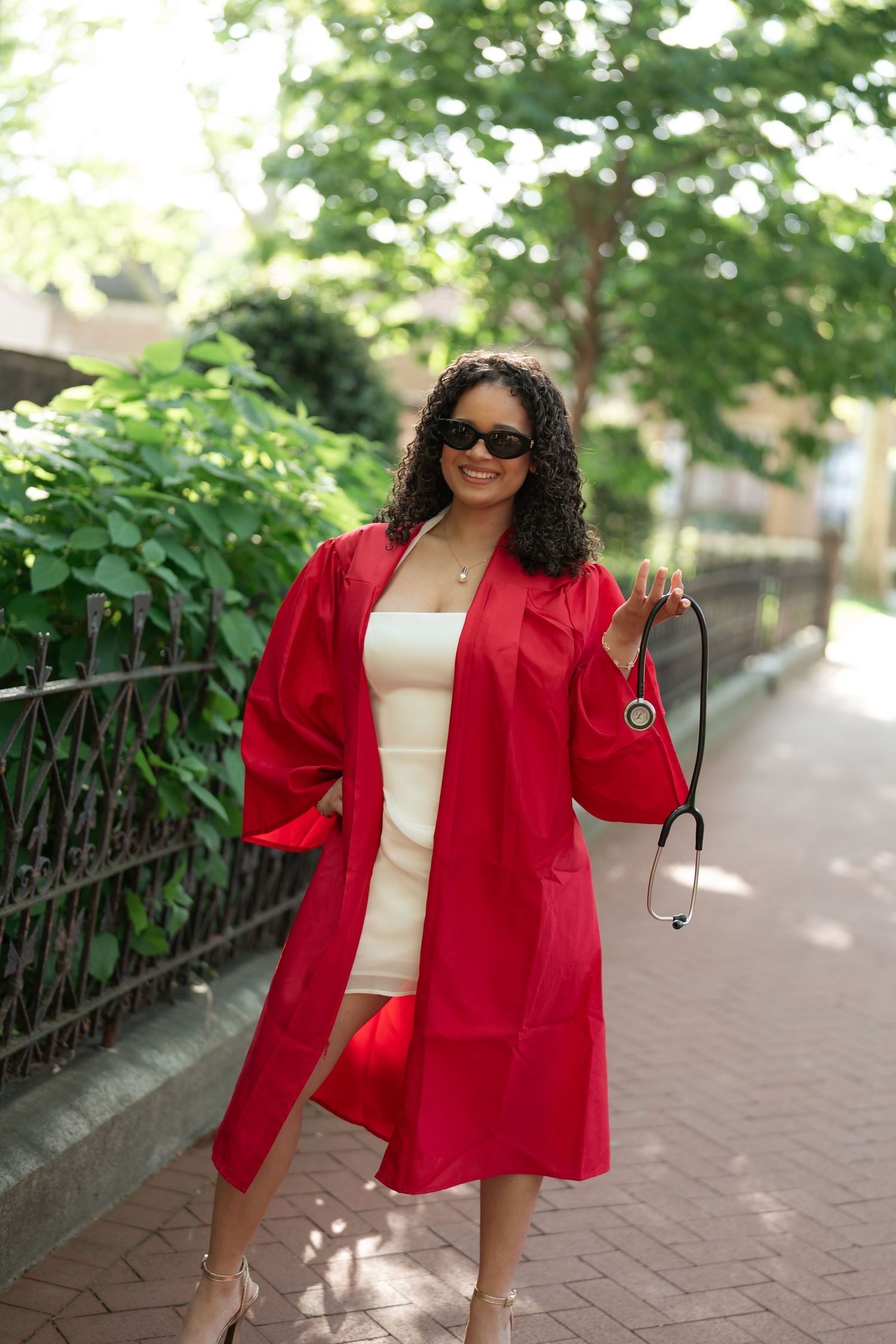 Smiling medical graduate in red gown holds stethoscope while walking on sunny tree-lined pathway.