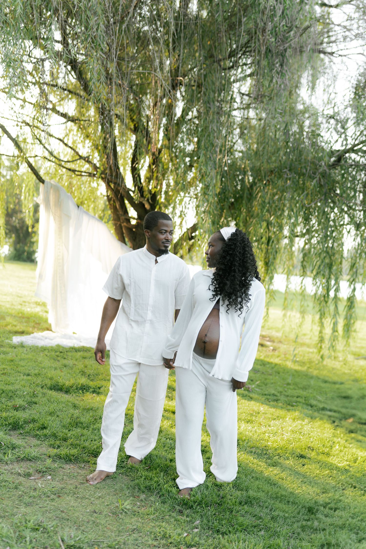 A couple in matching white outfits standing together under a willow tree on green grass.