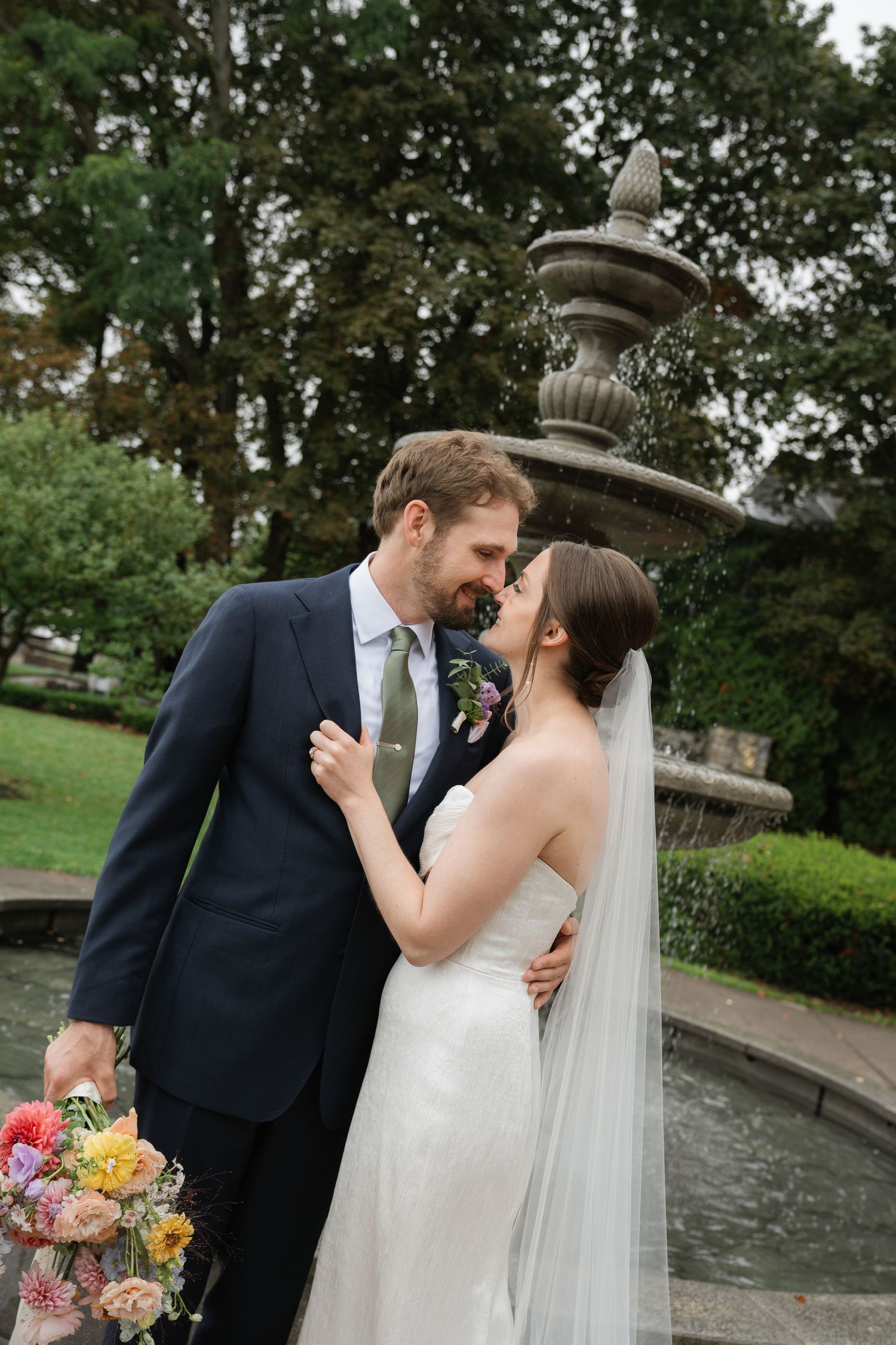 Romantic wedding couple sharing intimate kisses in an outdoor garden setting with a fountain in the background.