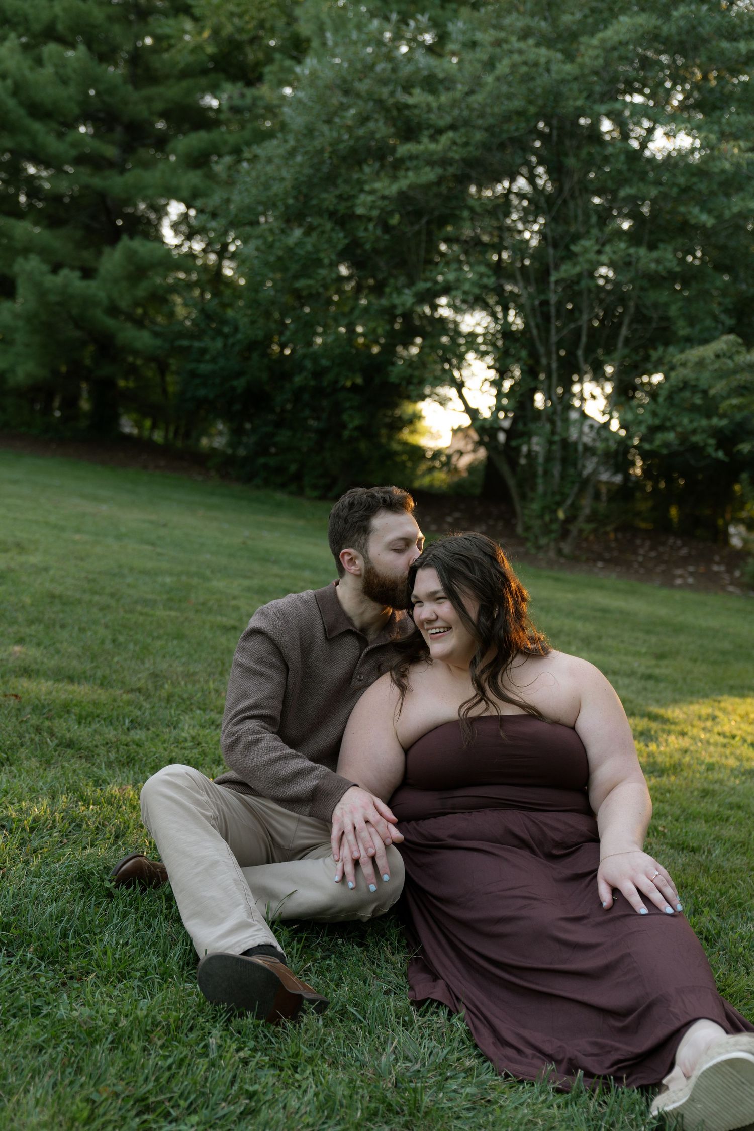 A romantic couple sits together on grass at sunset sharing an intimate moment in a park setting.