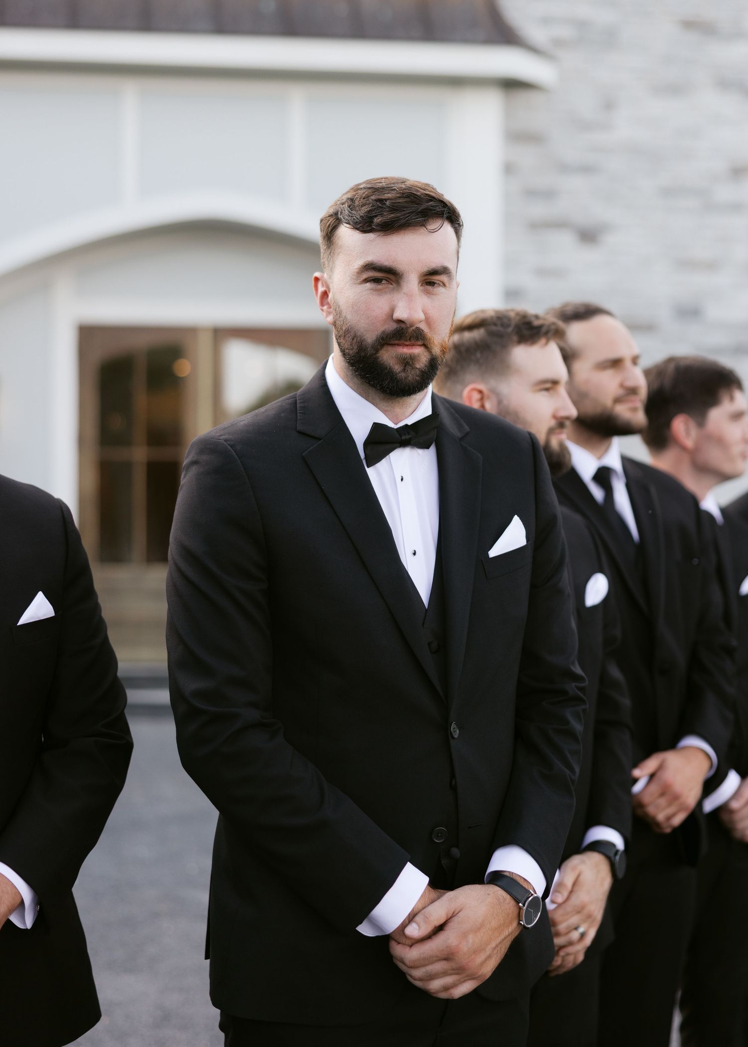 Handsome bearded groom in black tuxedo with bow tie stands with groomsmen at outdoor ceremony.