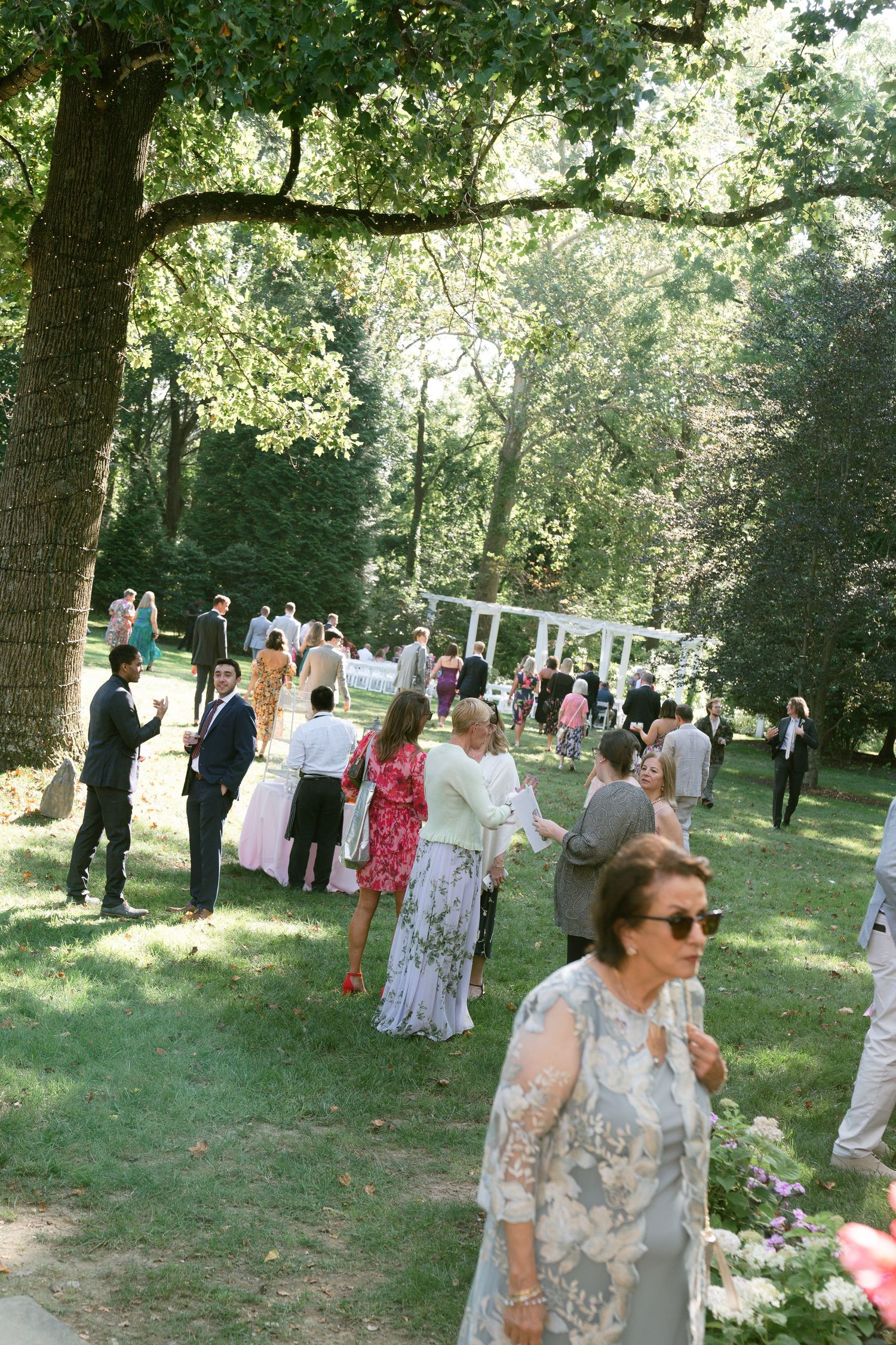 Guests mingling at an outdoor wedding reception under trees with white decor.