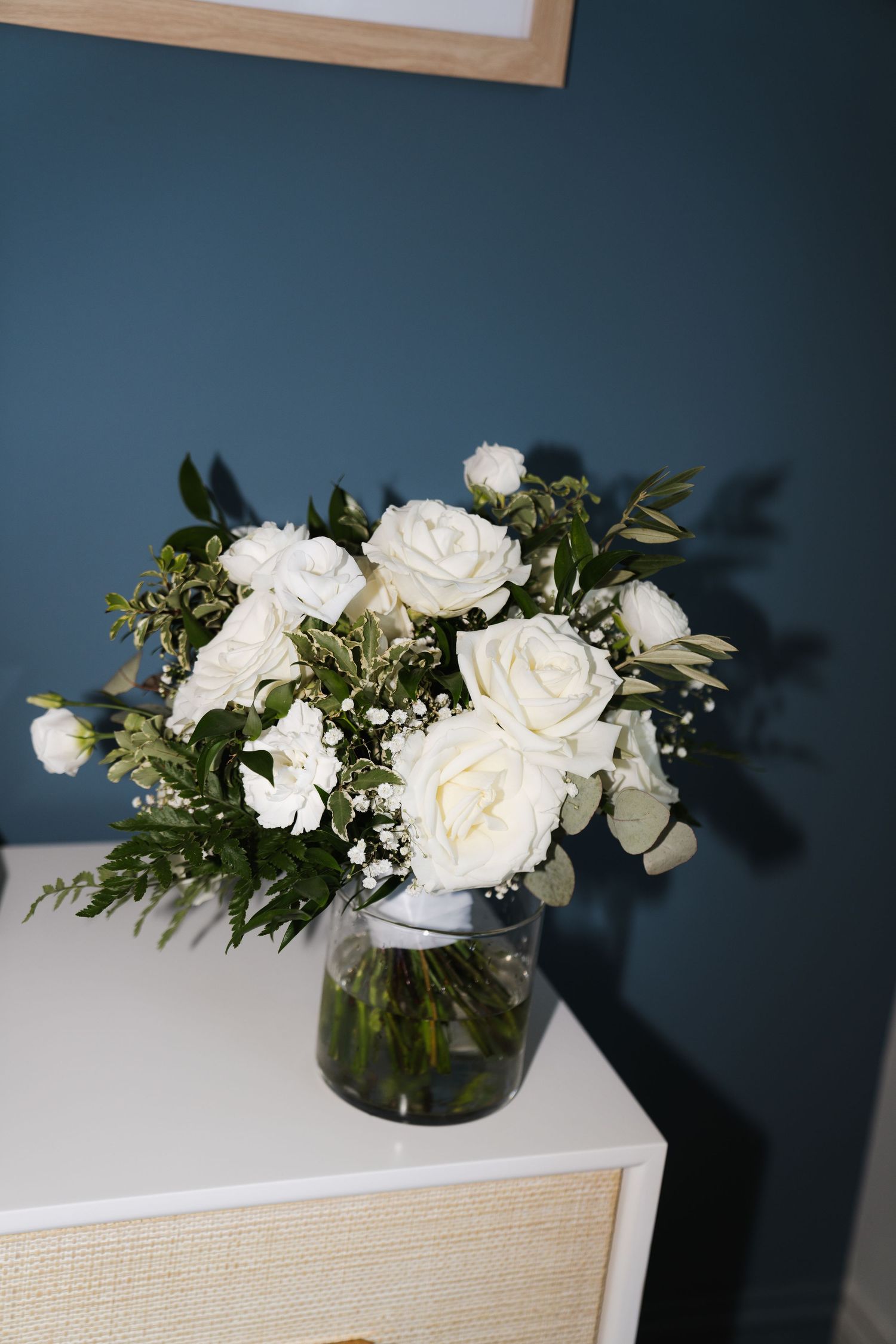 White roses and greenery arranged in clear glass vases against a dark blue wall create an elegant floral display.