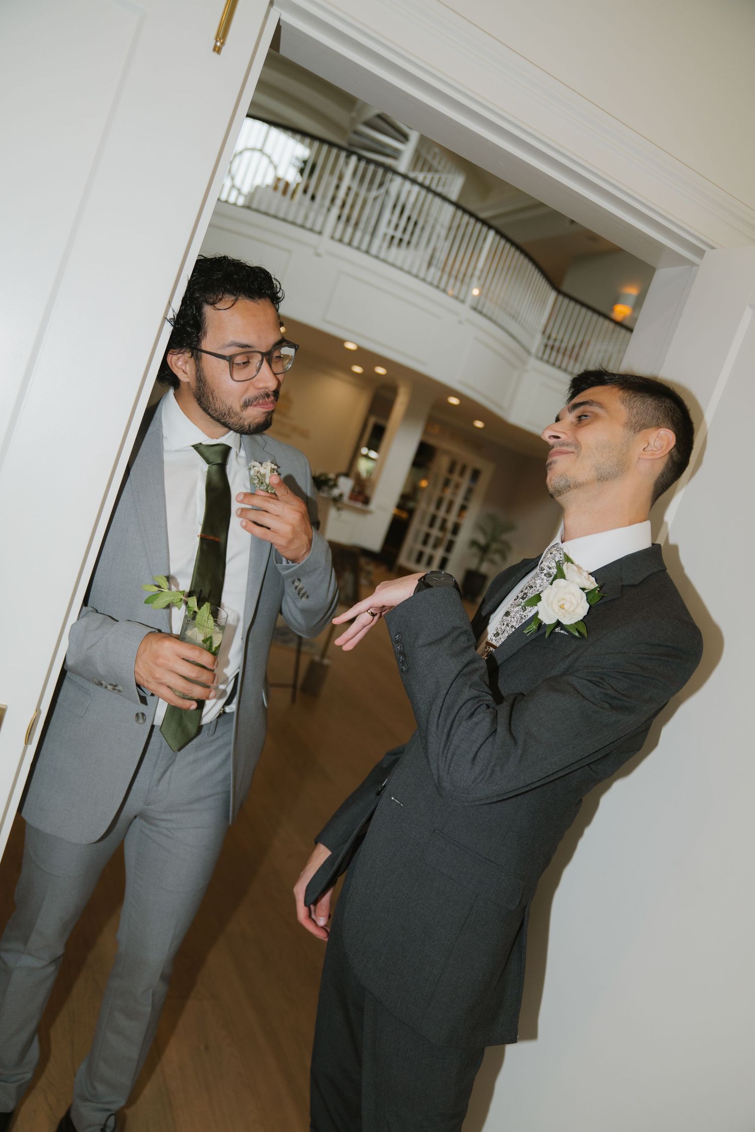 Two men in formal suits share a lighthearted moment in an elegant hallway with white walls and balcony above.