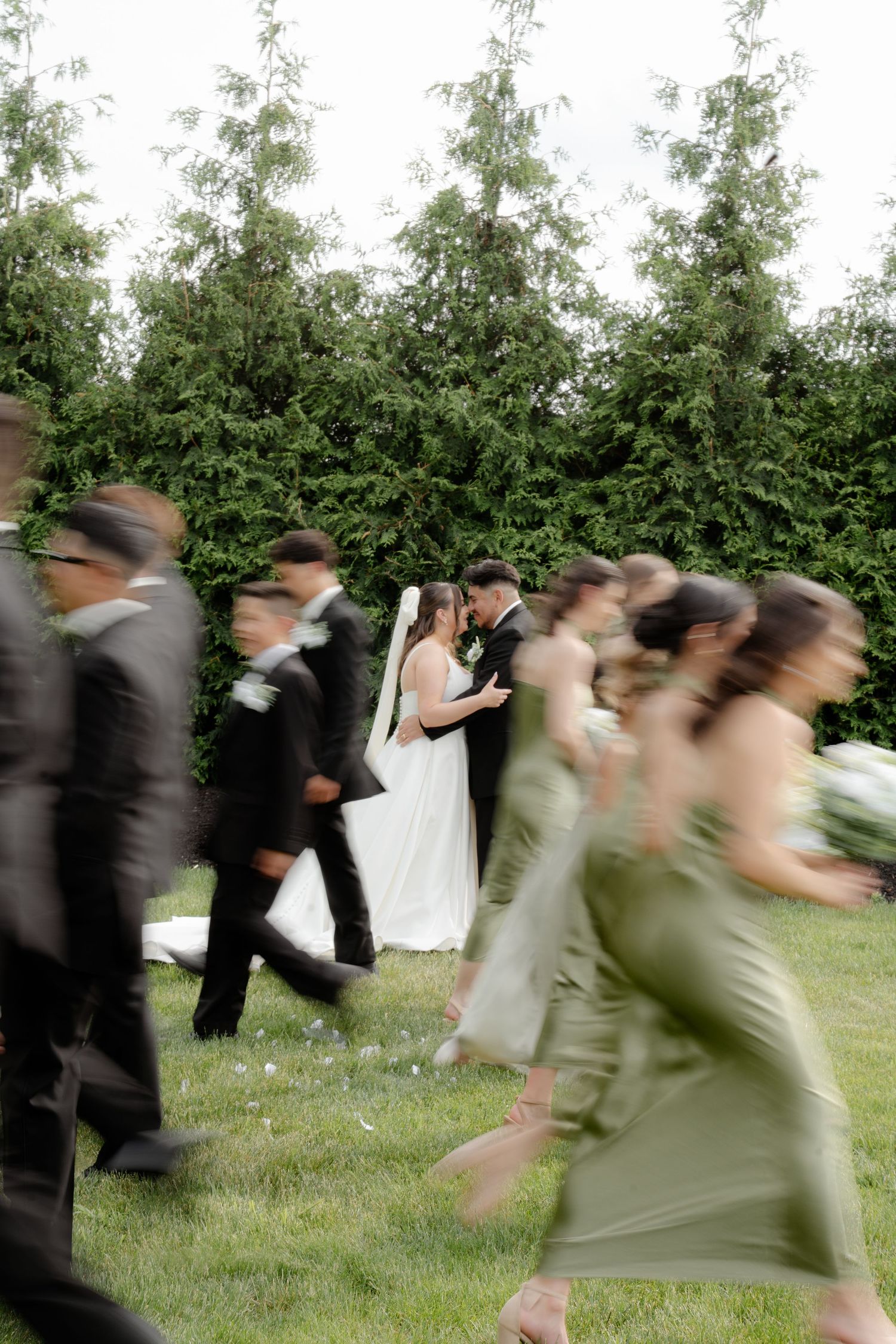 Motion-blurred sequence of wedding party running across a grassy lawn with trees in the background.