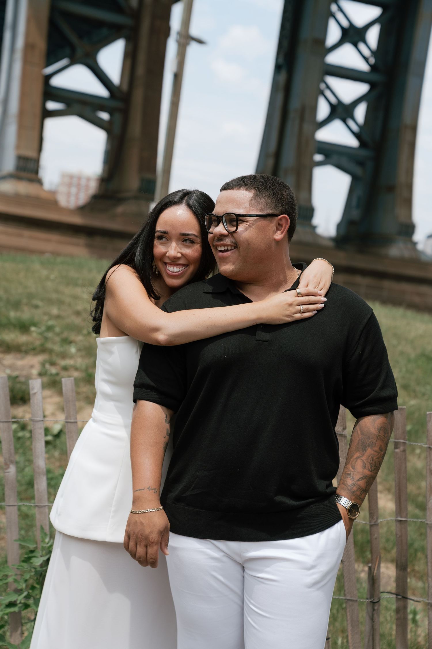 A smiling couple embraces outdoors under a large bridge, woman in white outfit hugging man in black shirt and white pants.