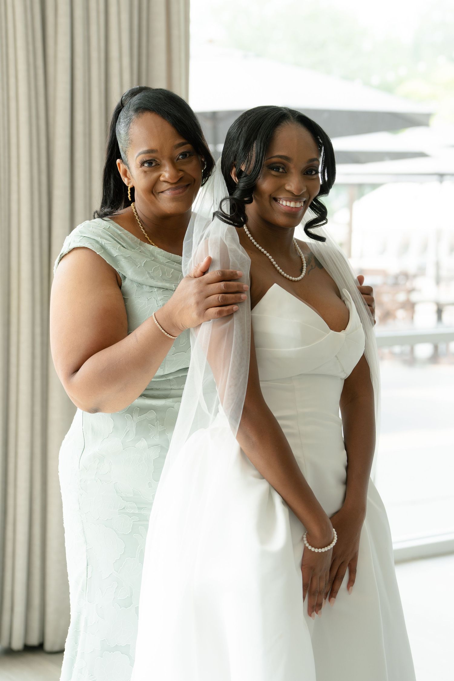 Bride in white wedding gown and veil smiles with woman in mint dress during wedding preparations.