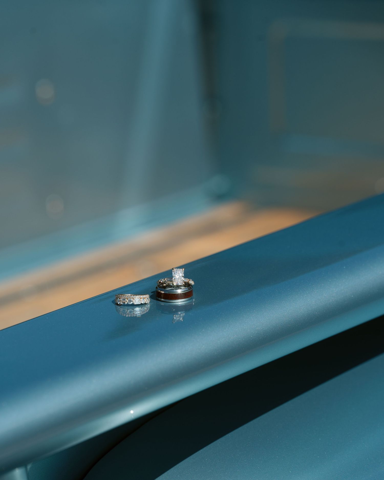 Two wedding rings, one diamond engagement ring and one wood inlay band, resting on a teal surface.