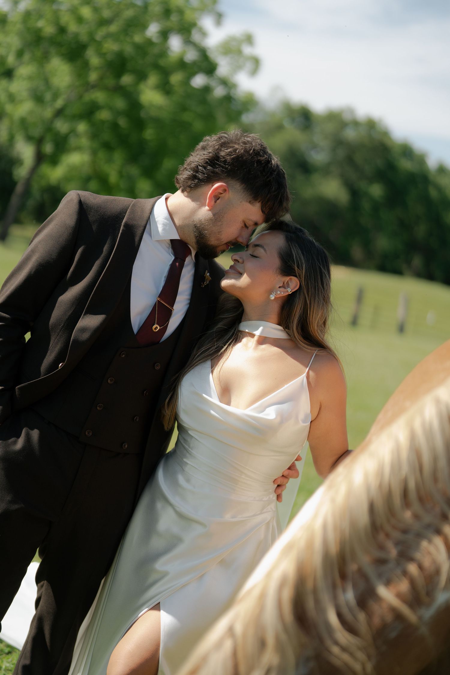 Bride and groom nearly kissing near a horse on a sunny farm with lush green trees behind.