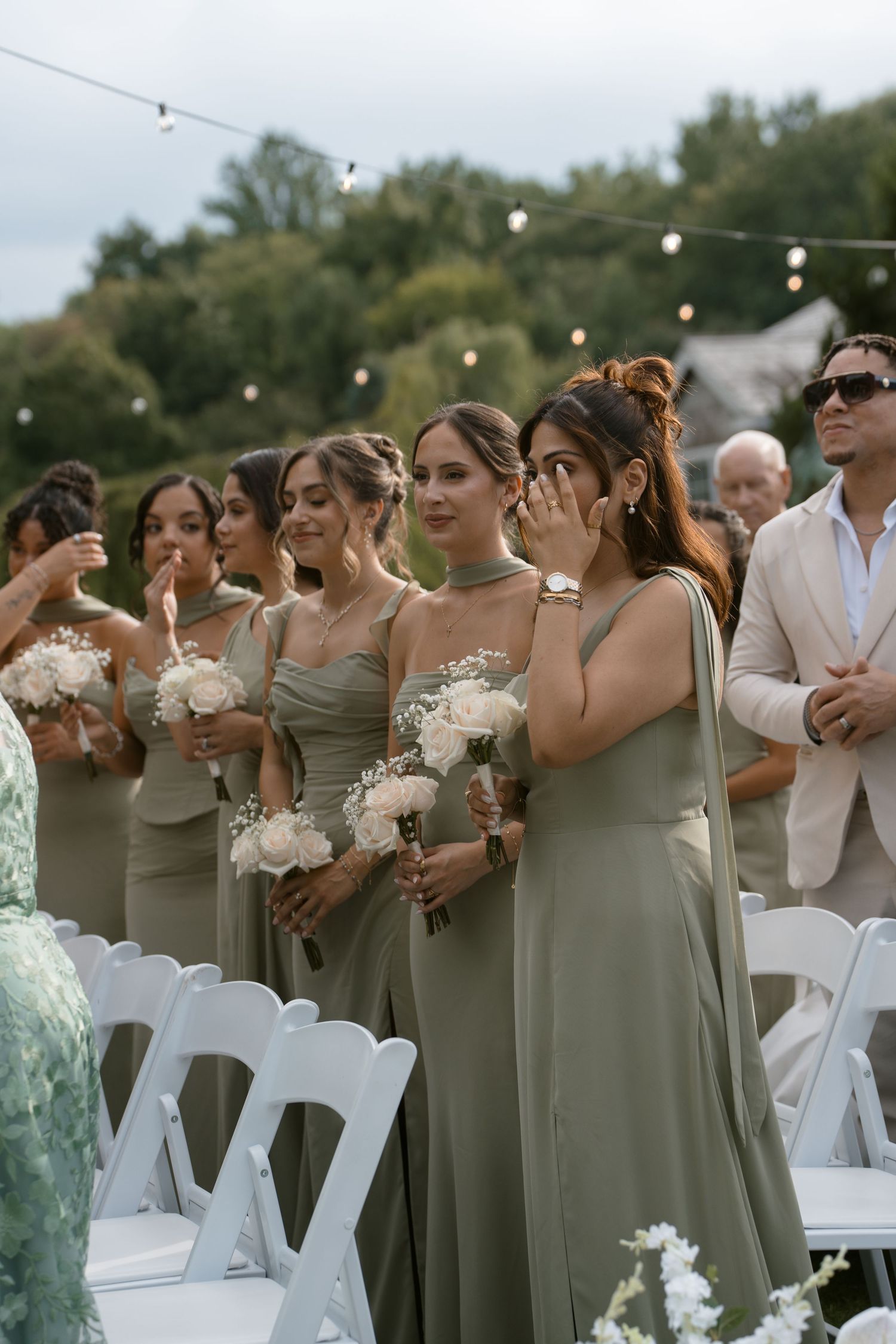Bridesmaids in sage green dresses holding white bouquets at an outdoor wedding ceremony, one wiping tears.