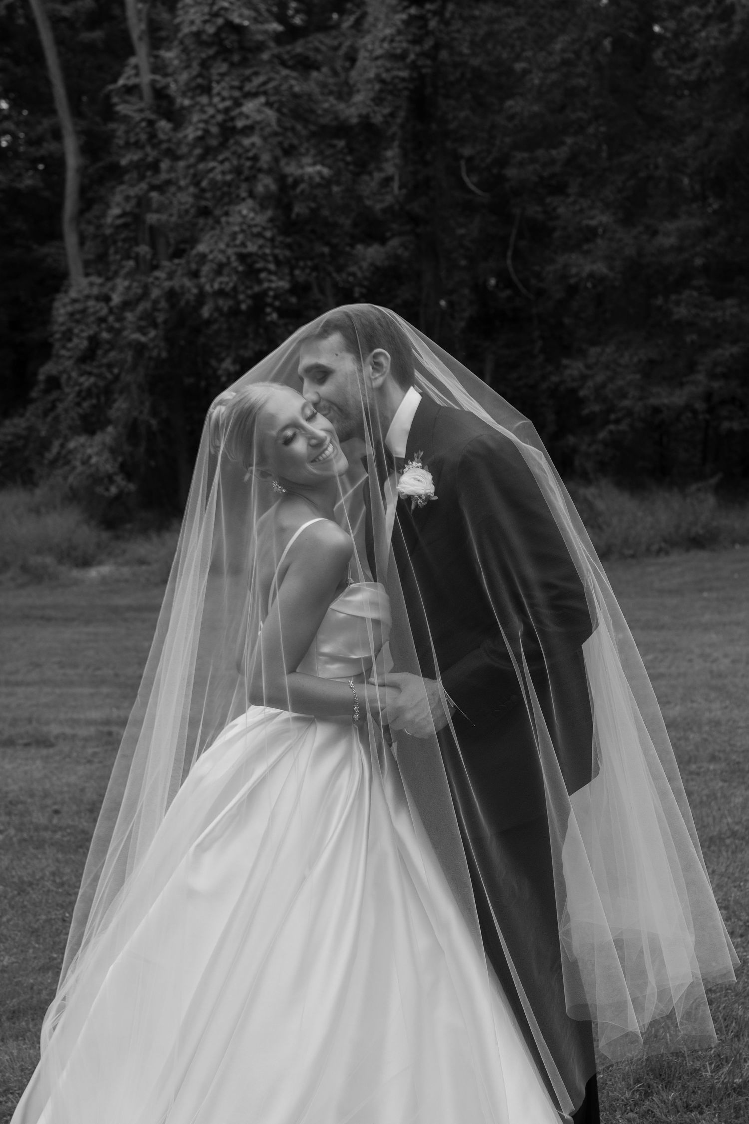 Bride and groom share romantic moment under wedding veil in black and white outdoor portrait.