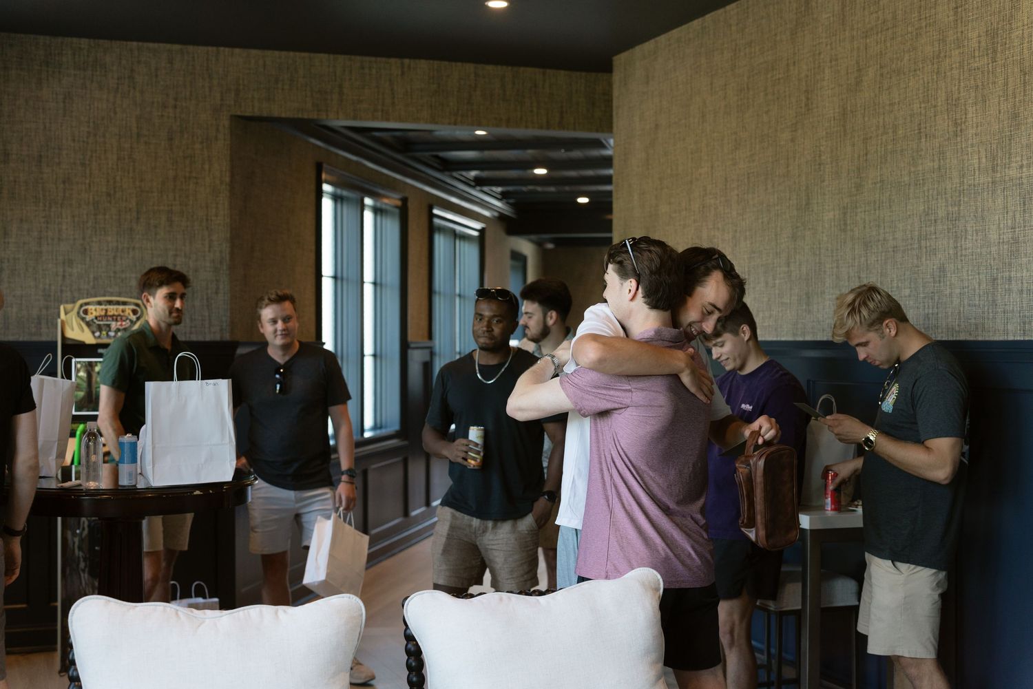 Group of young adults socializing indoors, two people hugging warmly while others stand nearby in a modern lounge setting.