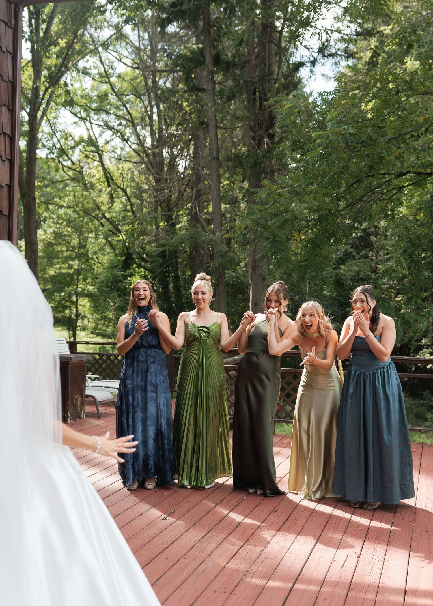 Bridesmaids in mismatched green and blue gowns react with joy on a wooden deck surrounded by lush trees.