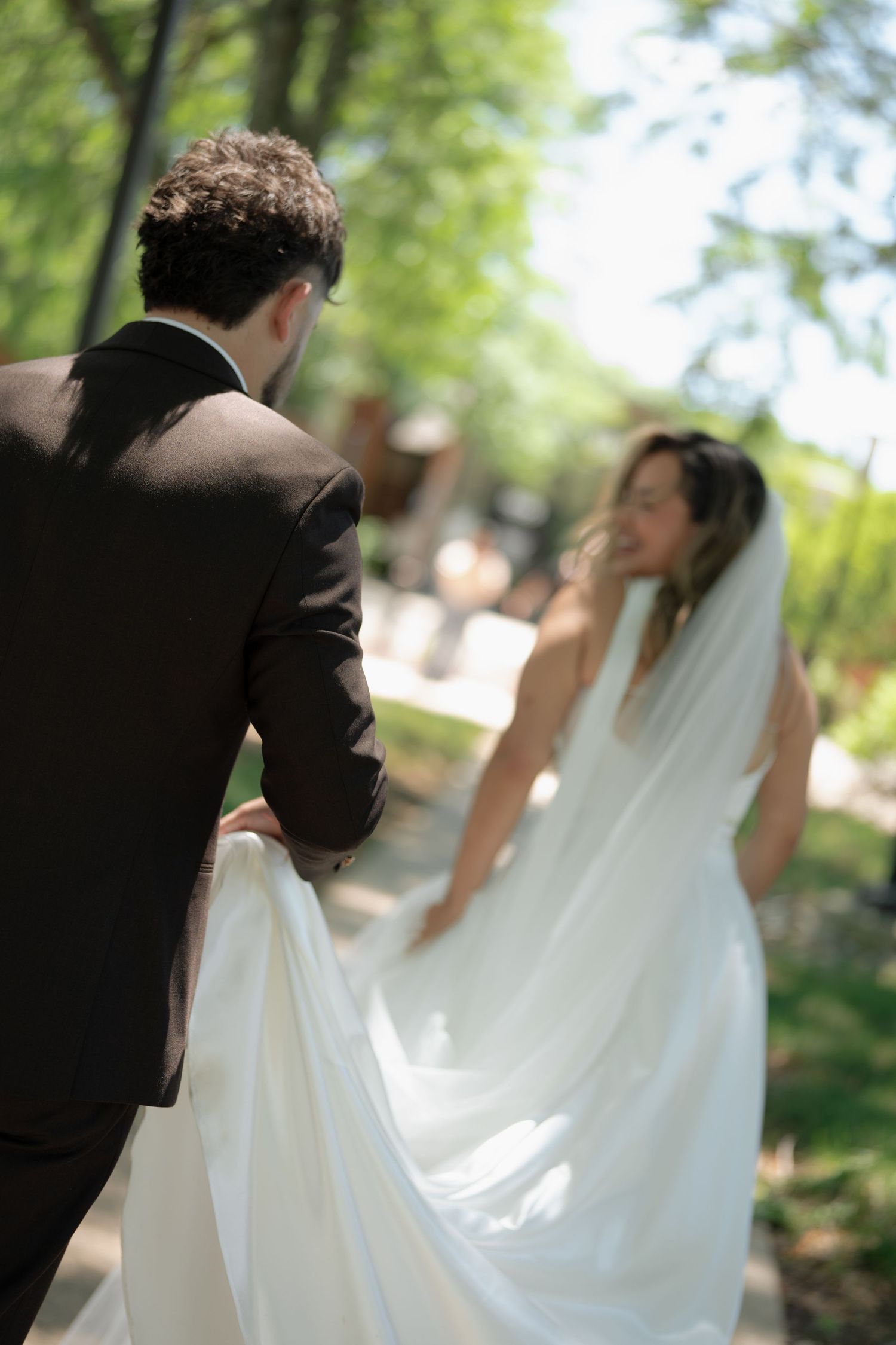 Groom in black suit holds bride's flowing white wedding dress train as she glances back outdoors.
