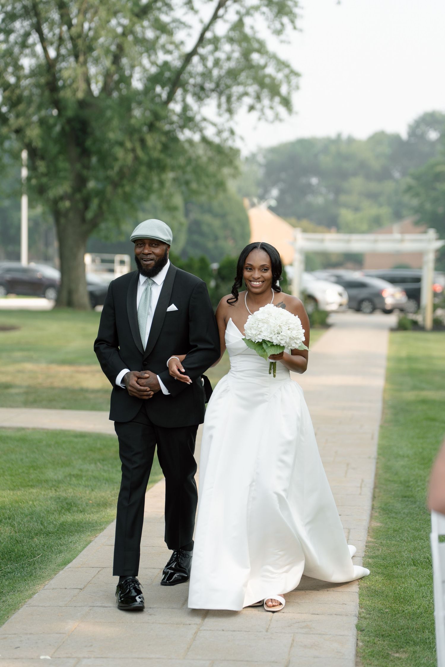 Smiling bride in white gown walks down aisle with escort in black suit and gray cap.