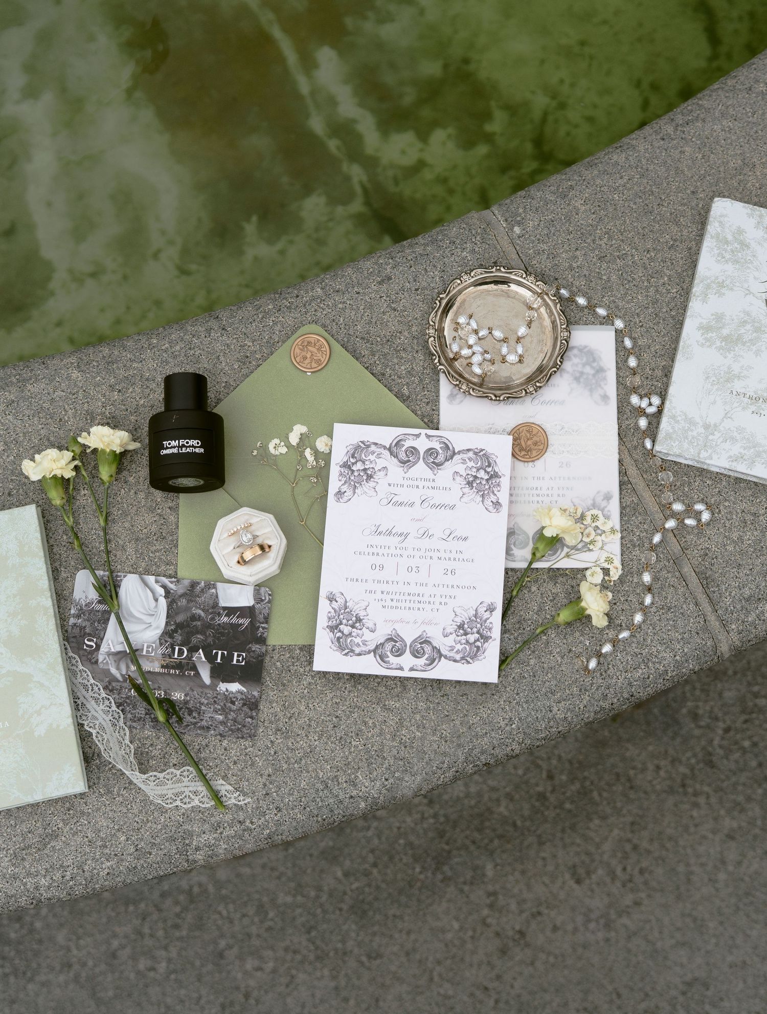 Elegant wedding invitation suite with florals, perfume, rings, and greenery on stone surface.