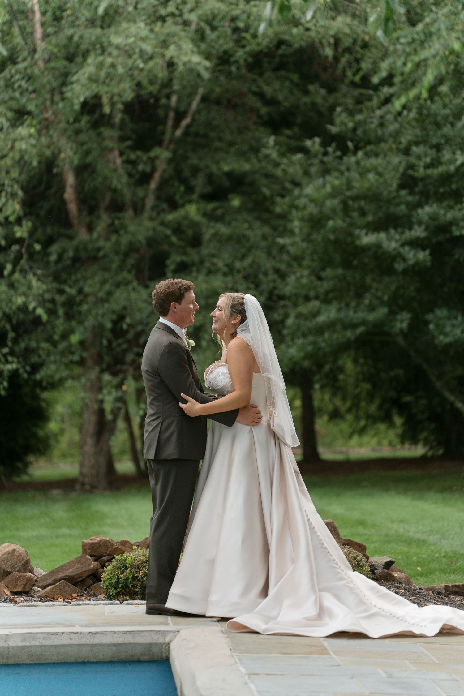Bride and groom share romantic moment by poolside surrounded by lush green trees in elegant wedding attire.