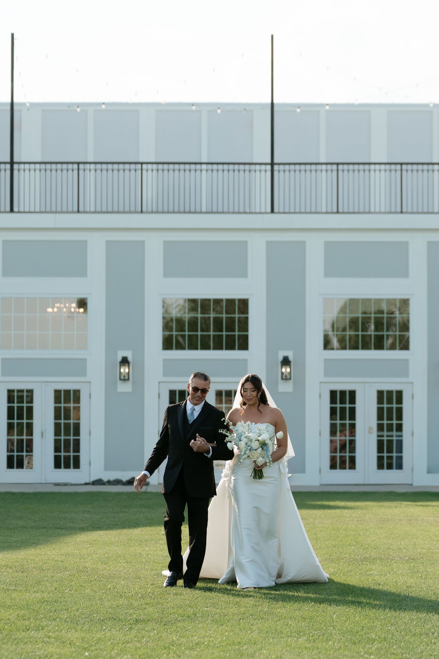 Father walks bride in white strapless gown across green lawn toward elegant white wedding venue.