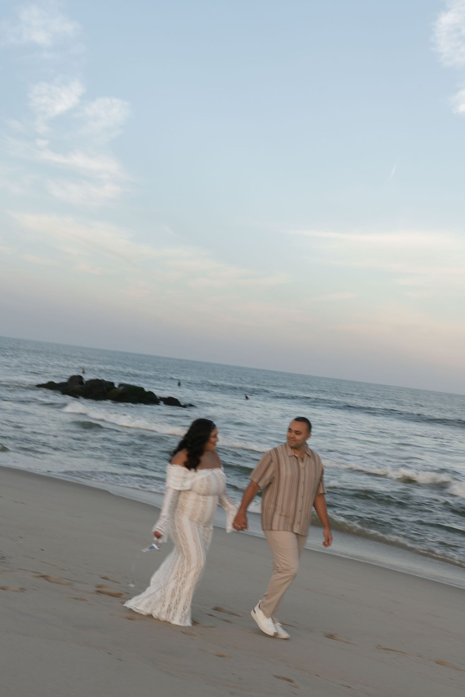 Couple holding hands walking on a sandy beach at sunset, woman in white dress, man in striped shirt and khakis.