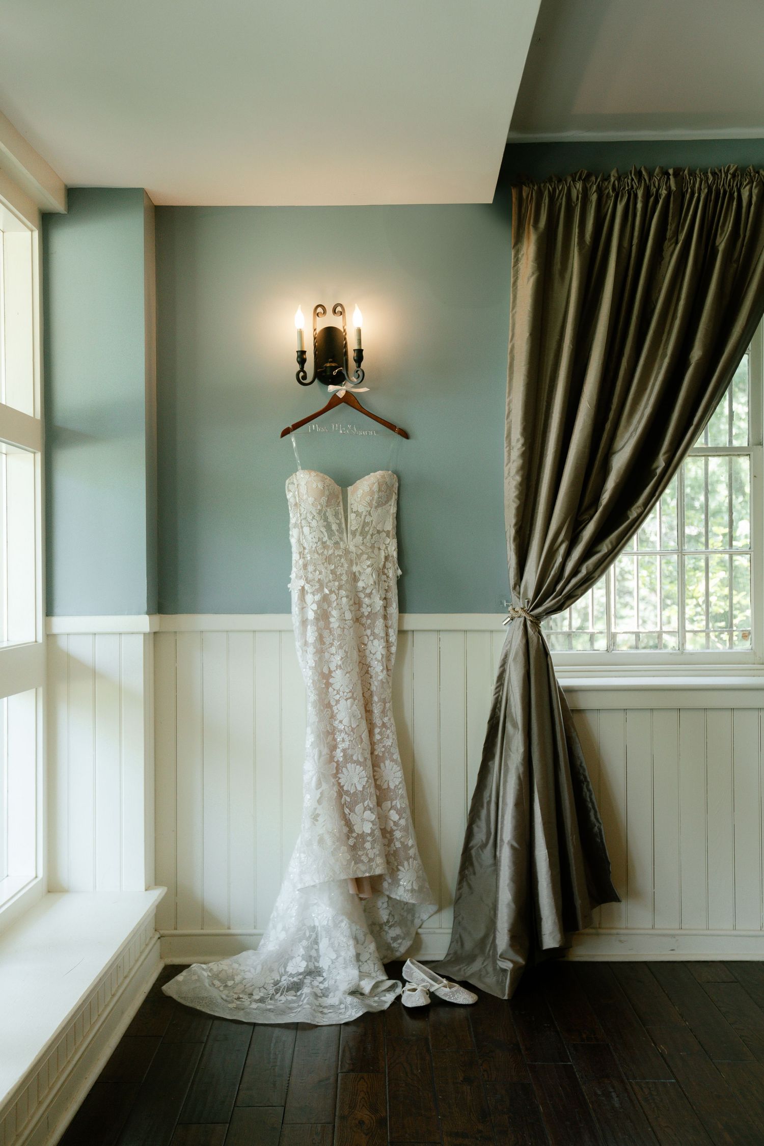 A white lace wedding dress hangs elegantly against dark curtains in a room with hardwood floors.