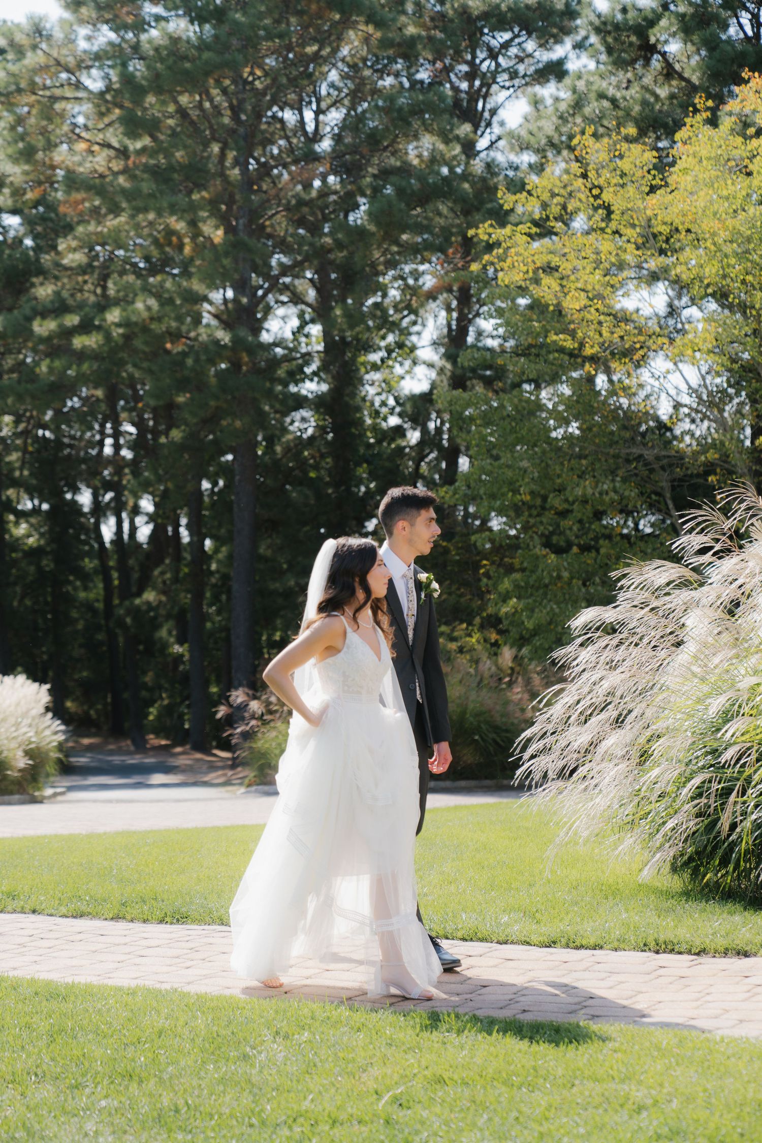 A couple in wedding attire walk together on a paved garden path surrounded by lush greenery and trees on a sunny day.