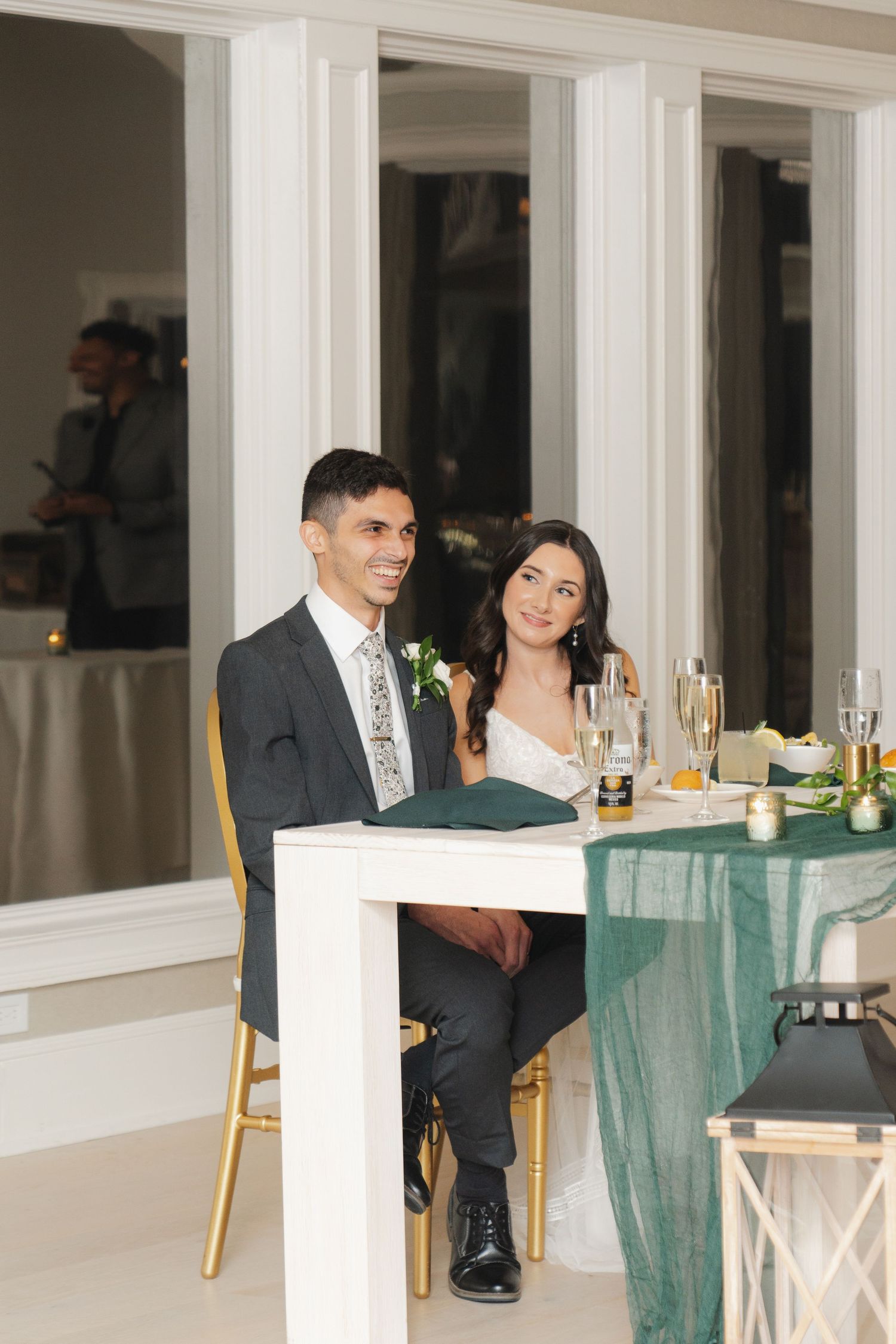 A couple sits at an elegant reception table with champagne glasses and emerald green decor at a formal event.