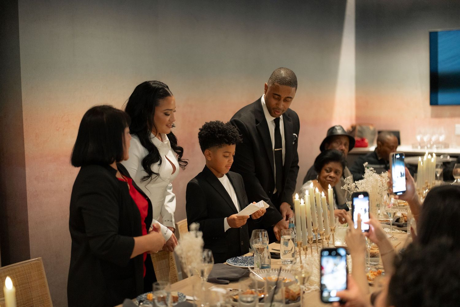 Wedding guests gather around a formal dinner table with phones capturing the moment.
