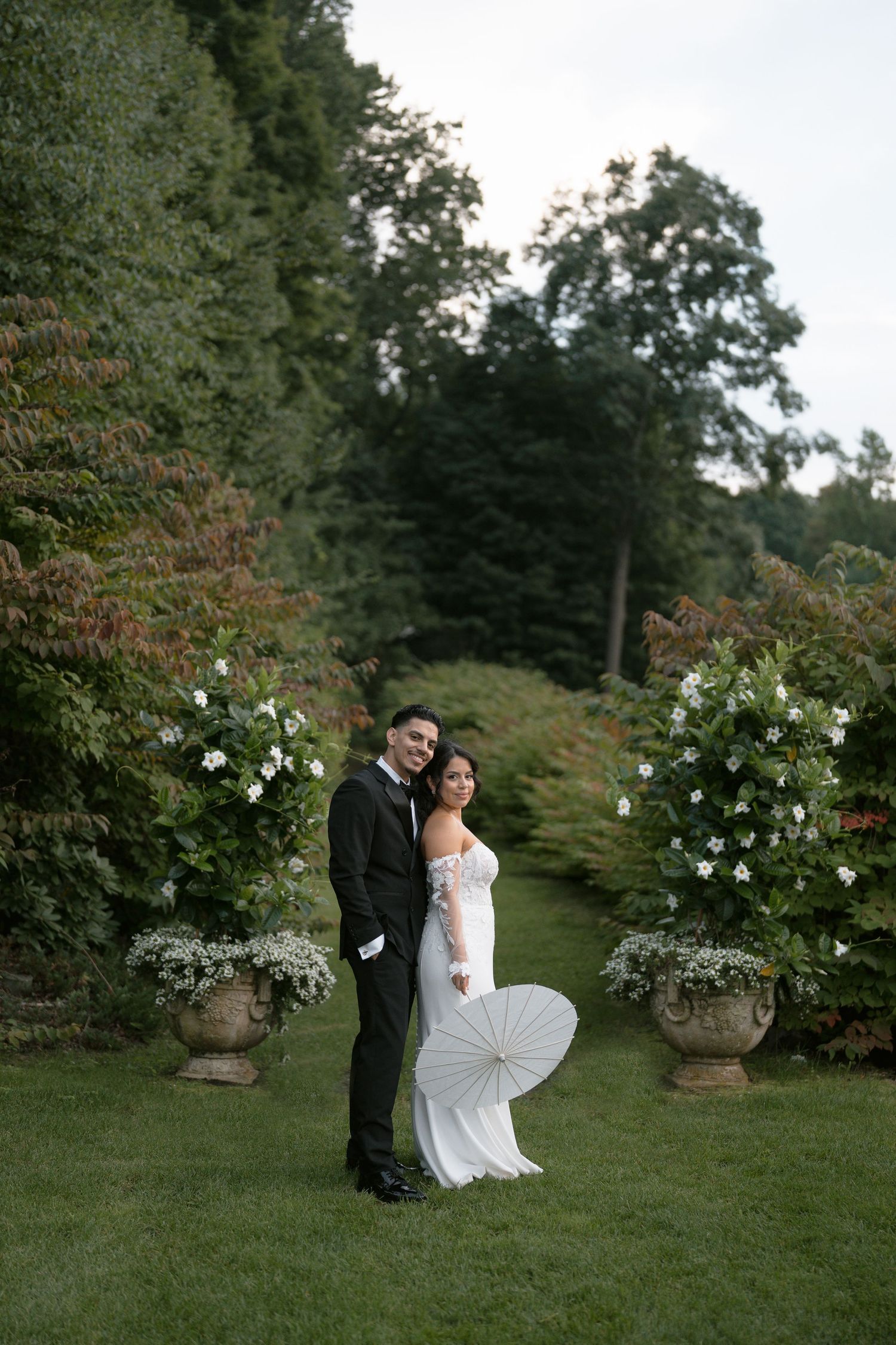 Bride and groom posing in lush garden with white floral arrangements, bride holding parasol in fitted wedding gown.