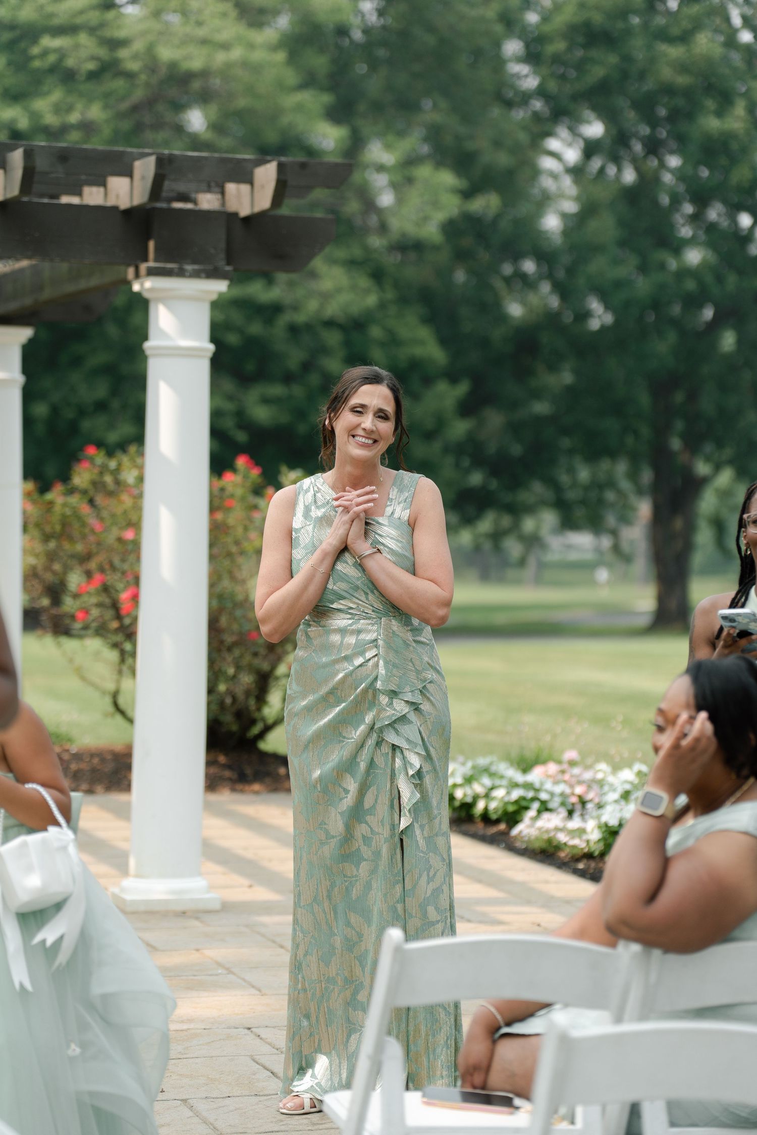 Woman in sage green floral dress smiling at outdoor wedding ceremony beside white pergola and garden.