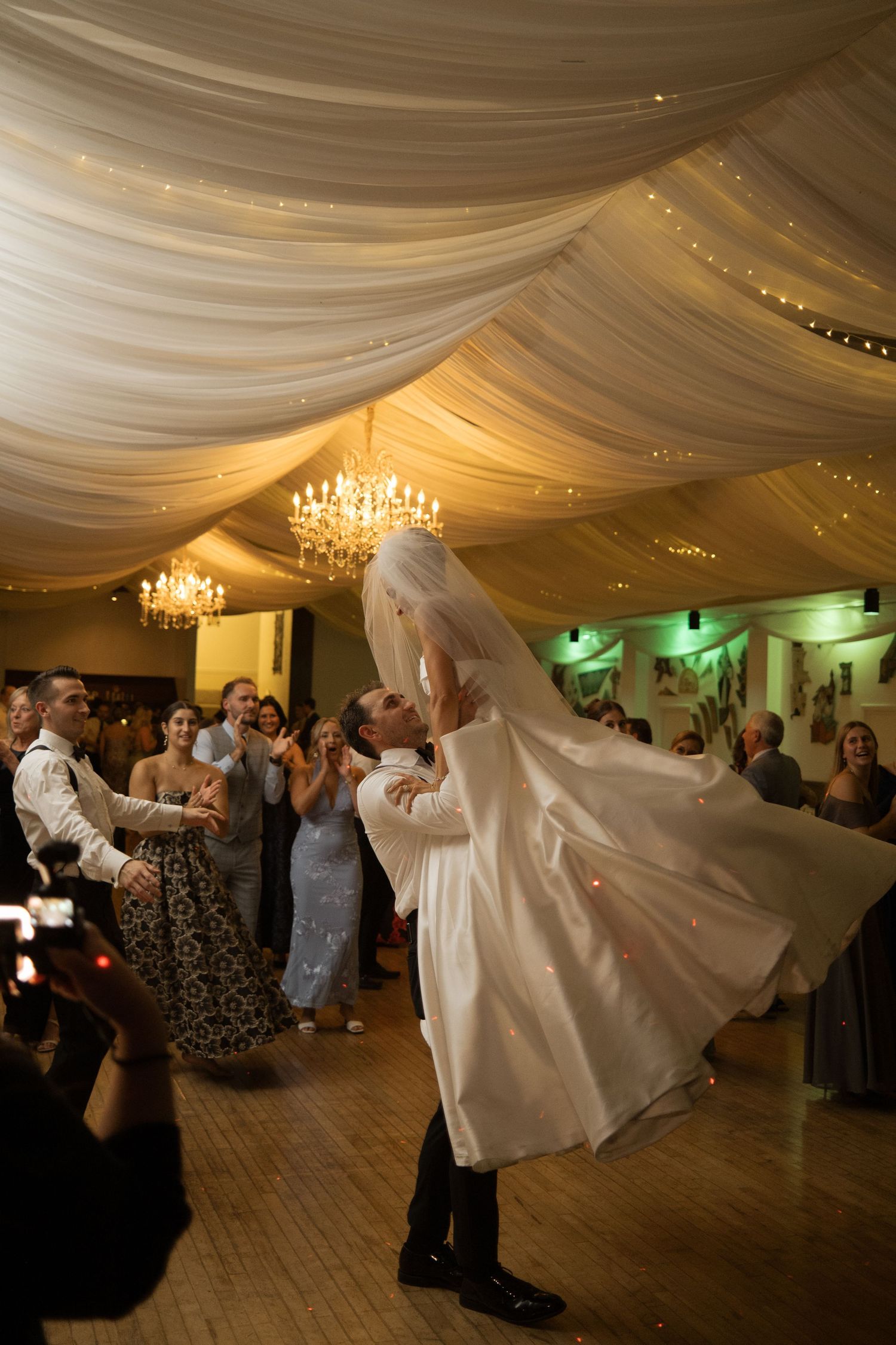 Groom lifts bride in white wedding dress during first dance at elegant reception with draped ceiling and chandeliers.