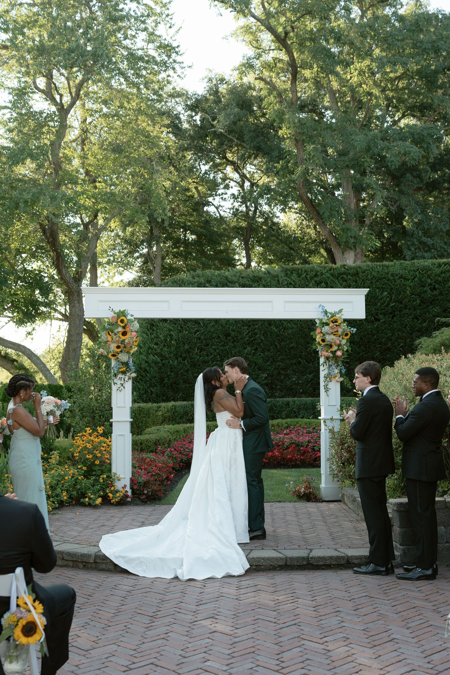 Bride and groom share first kiss under white floral arch surrounded by colorful garden at outdoor ceremony.