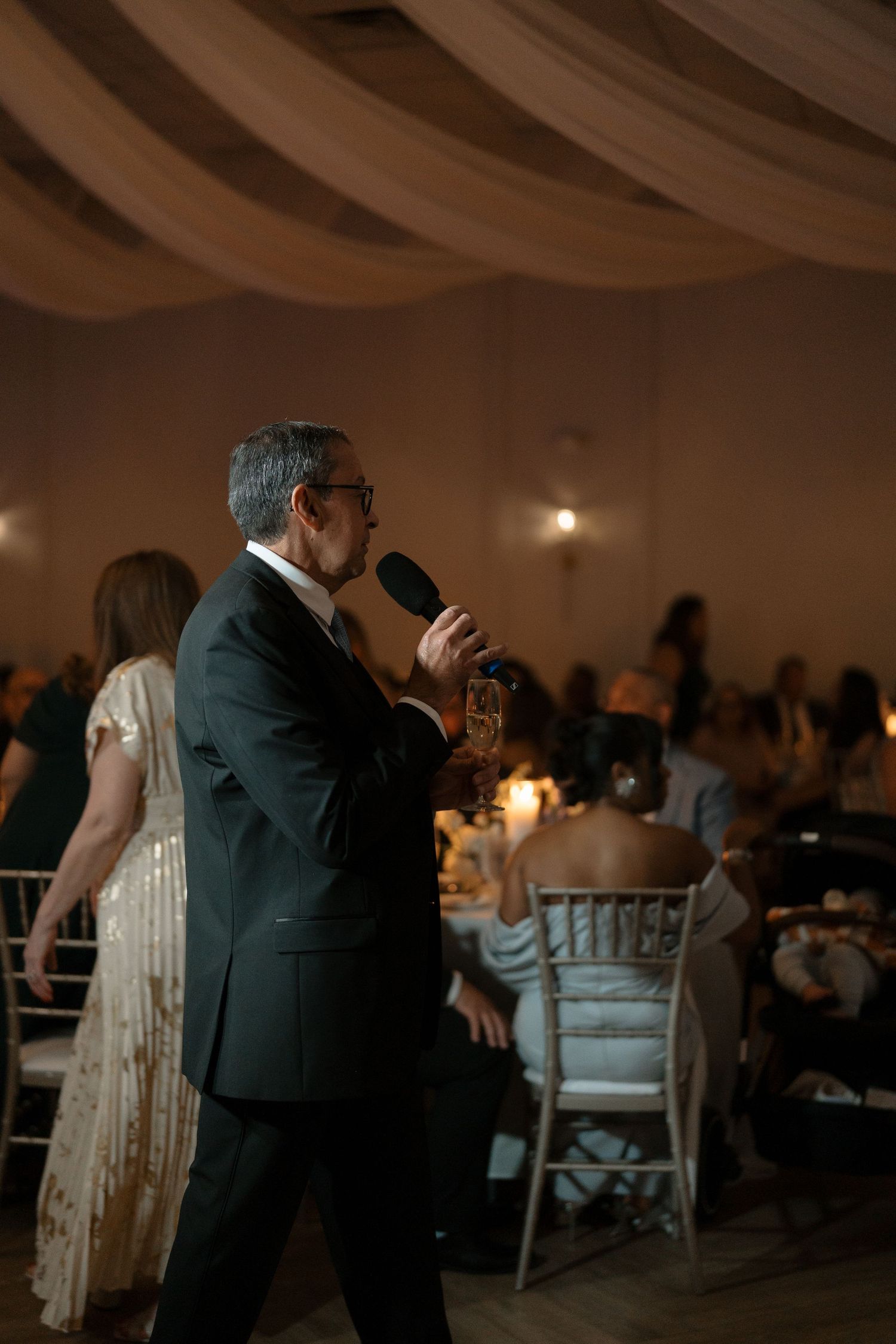 Man in dark suit gives a toast with microphone at a candlelit wedding reception dinner.