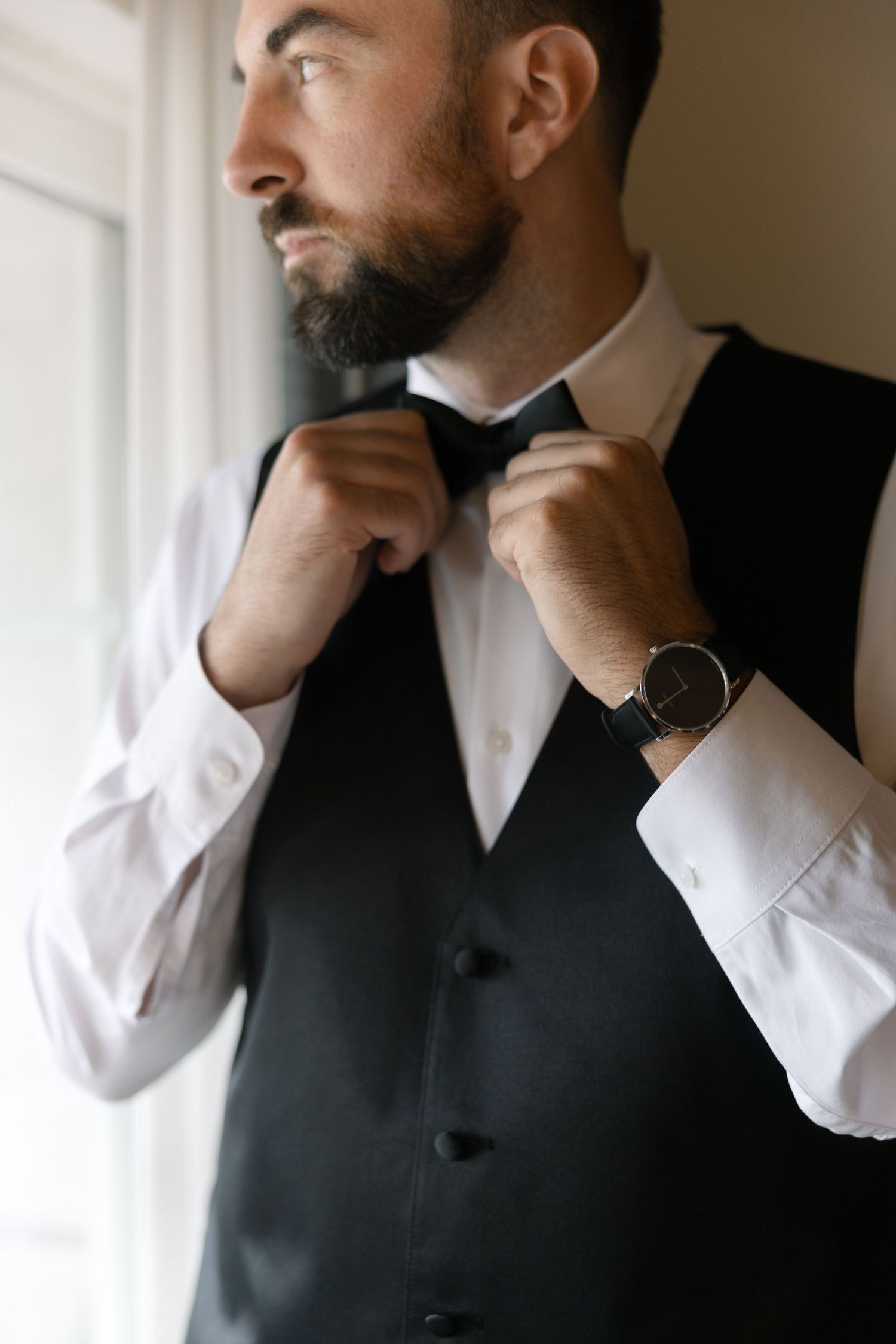 Bearded groom in black vest and white shirt adjusting bow tie while wearing elegant watch before wedding.